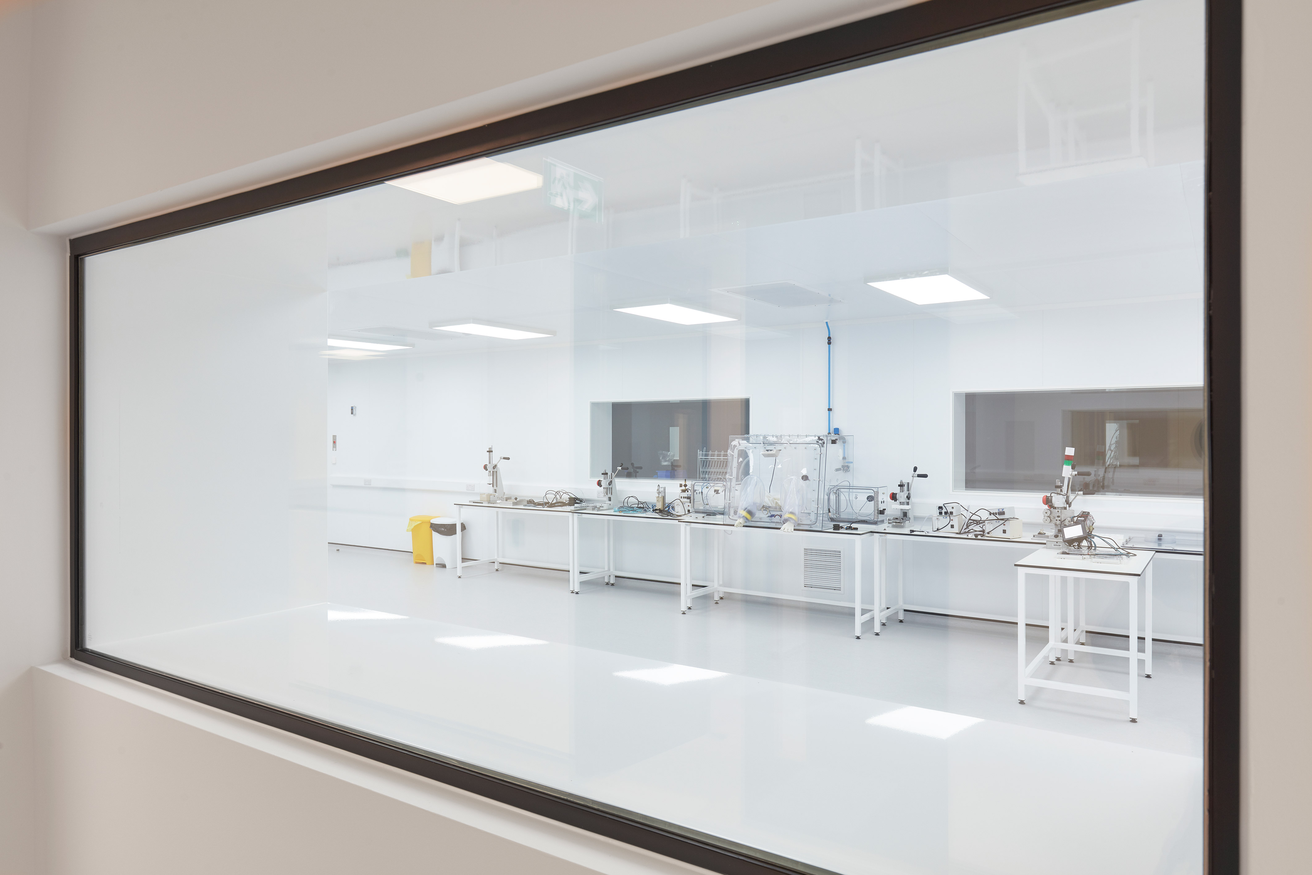 View through a large interior window into a bright, sterile cleanroom with white floors, workstations, and specialized laboratory equipment.