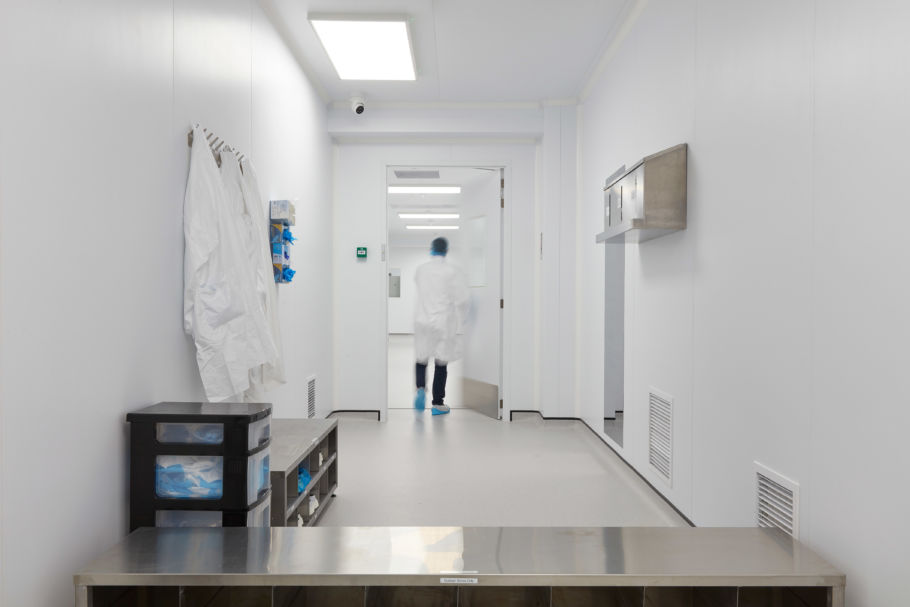 Sterile laboratory corridor with white walls and flooring, lab coats hanging on the wall, storage shelves, and a person in protective clothing walking through a doorway.