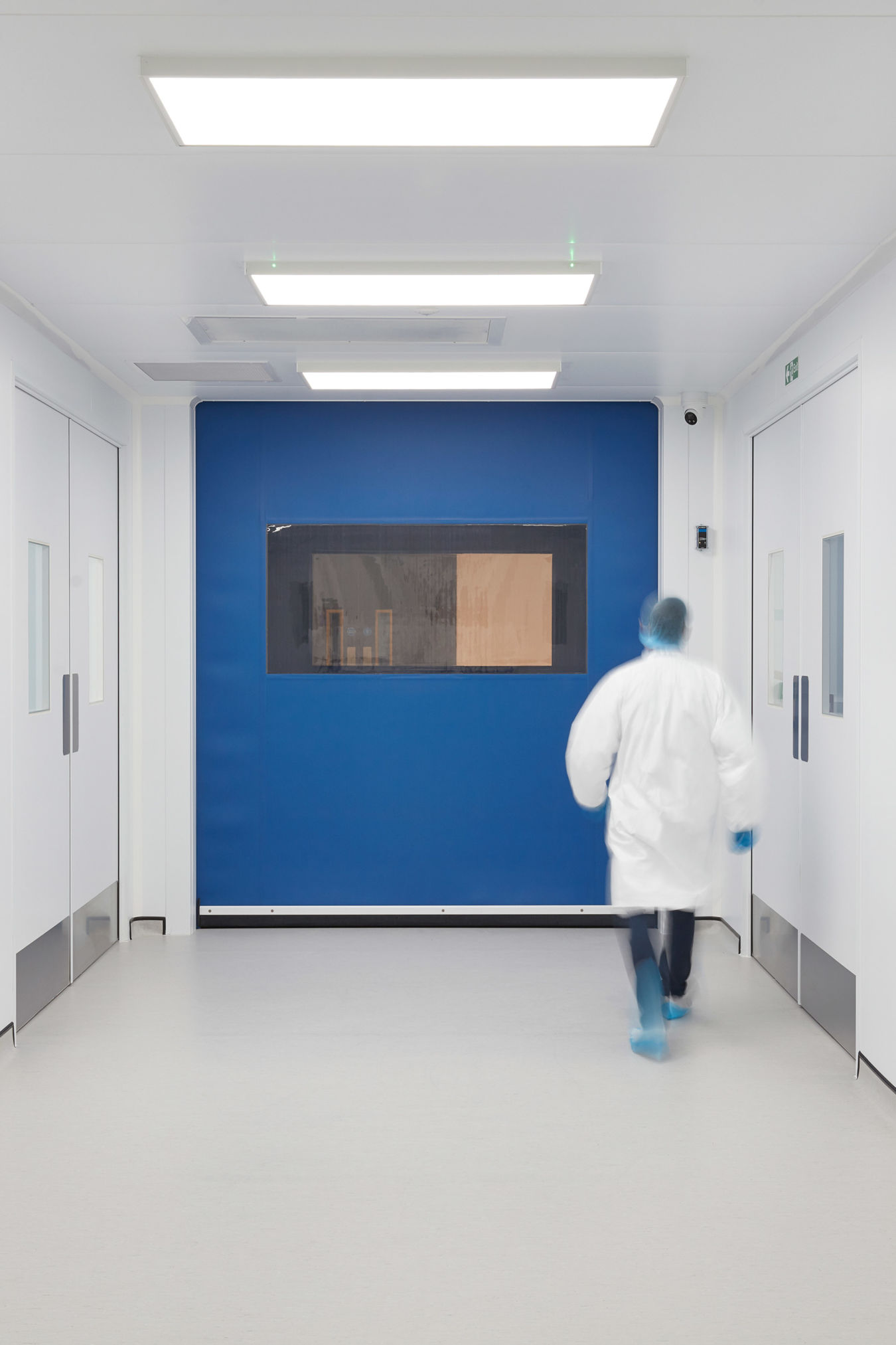 Person in a white lab coat walks down a sterile, brightly-lit corridor with white walls and doors, blue section, and a caution sign reading "Danger - Risk of Infection."