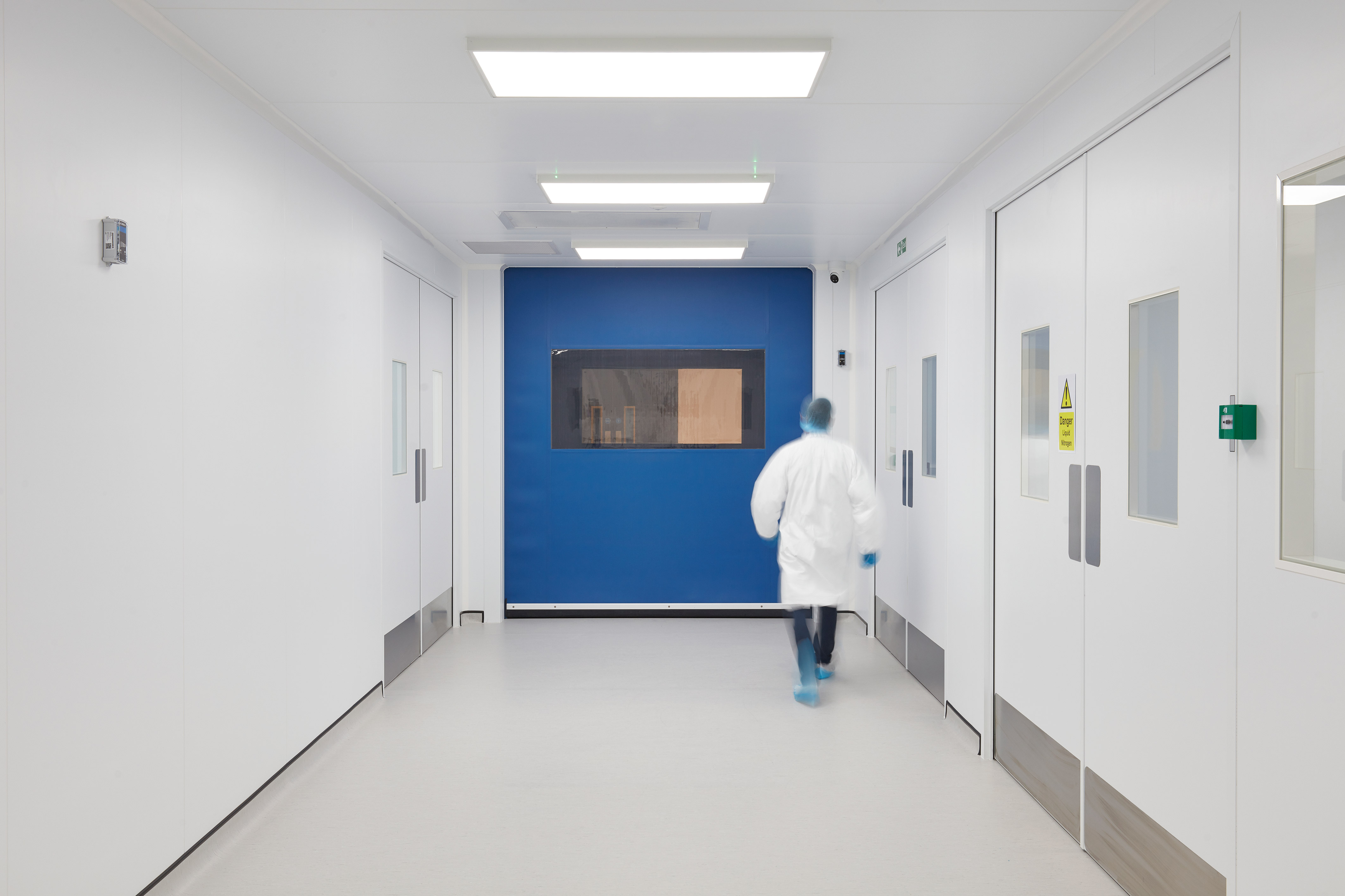 Person in a white lab coat walks down a sterile, brightly-lit corridor with white walls and doors, blue section, and a caution sign reading "Danger - Risk of Infection."