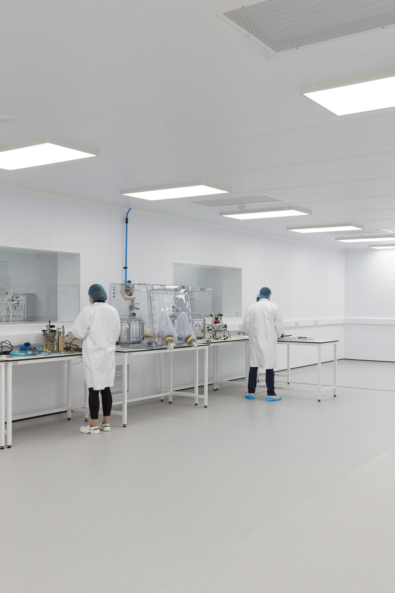 Spacious cleanroom environment with bright lighting, white walls, lab benches, and two technicians in lab coats and protective gear working at separate stations.