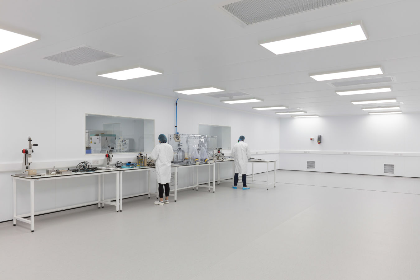Spacious cleanroom environment with bright lighting, white walls, lab benches, and two technicians in lab coats and protective gear working at separate stations.