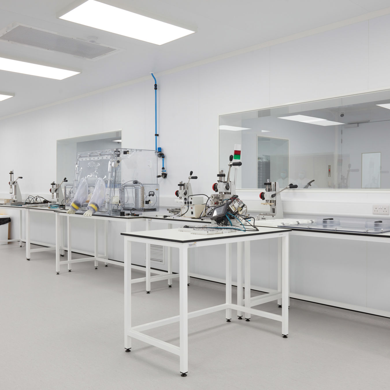 Clean, modern laboratory with white walls and bright lighting. Neat benches hold scientific equipment, including a glove box and various tools. The room is orderly and sterile