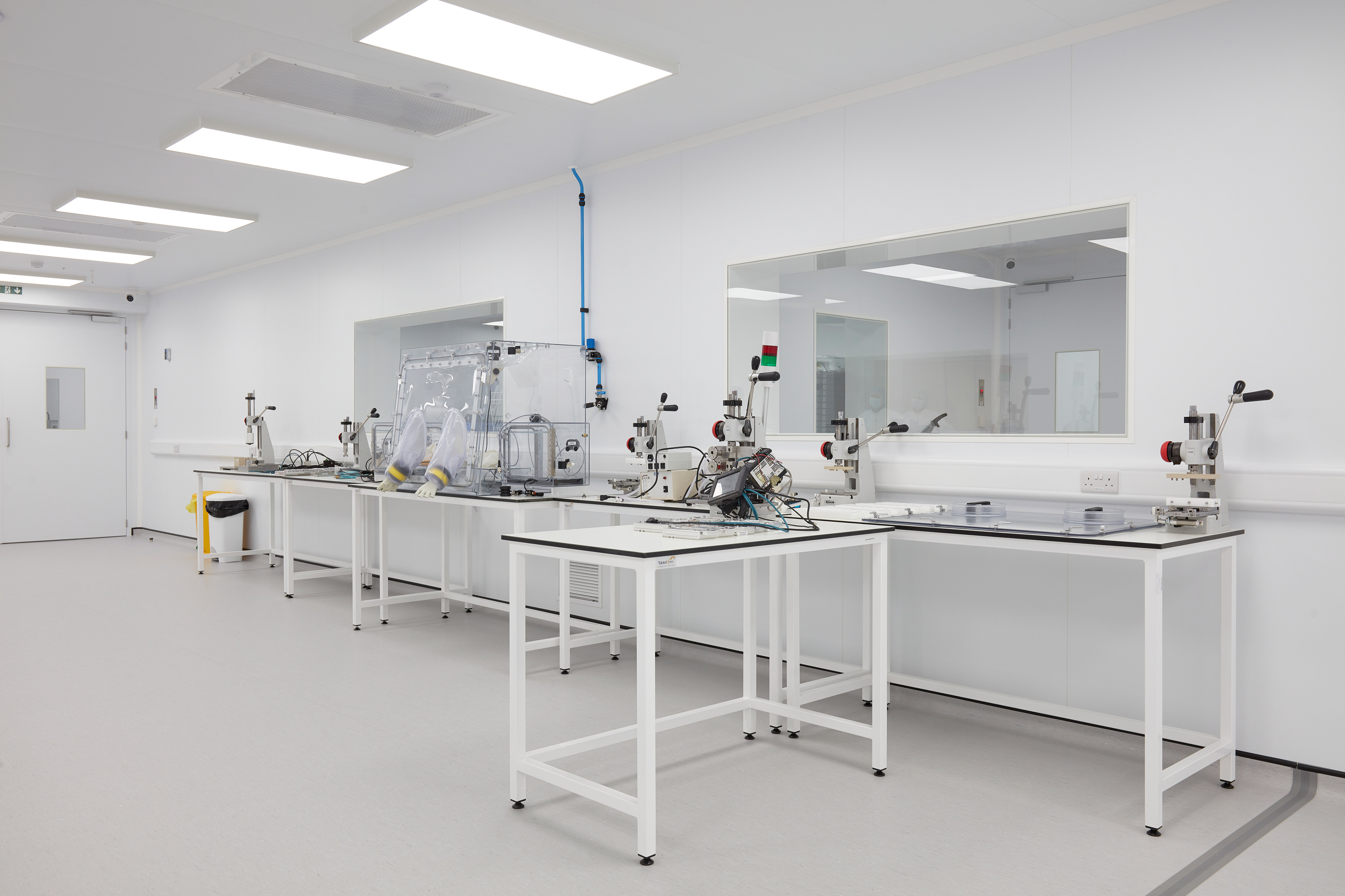 Clean, modern laboratory with white walls and bright lighting. Neat benches hold scientific equipment, including a glove box and various tools. The room is orderly and sterile