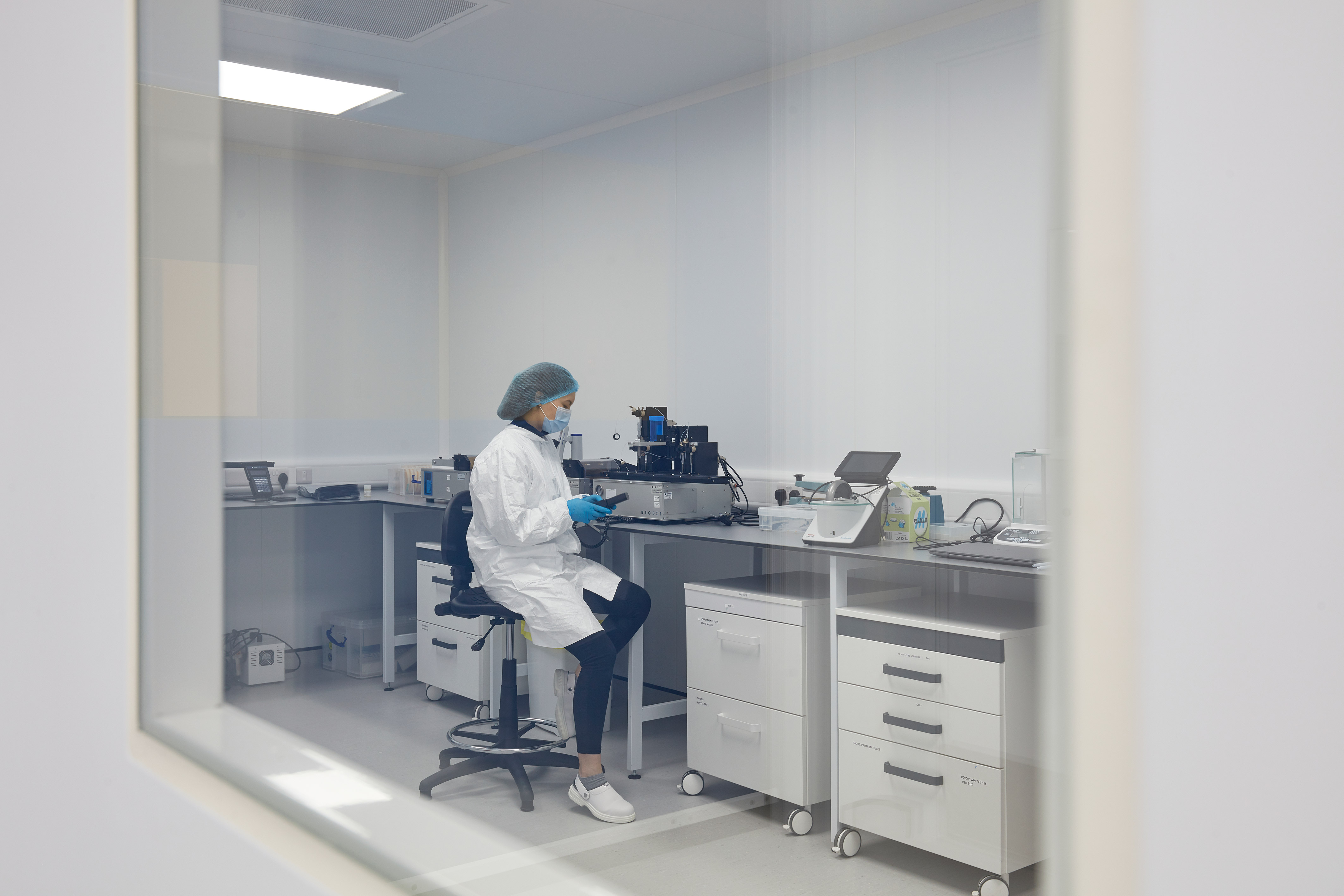 scientist in a lab coat and gloves works alone at a lab bench, surrounded by equipment and storage. The scene is calm and methodical