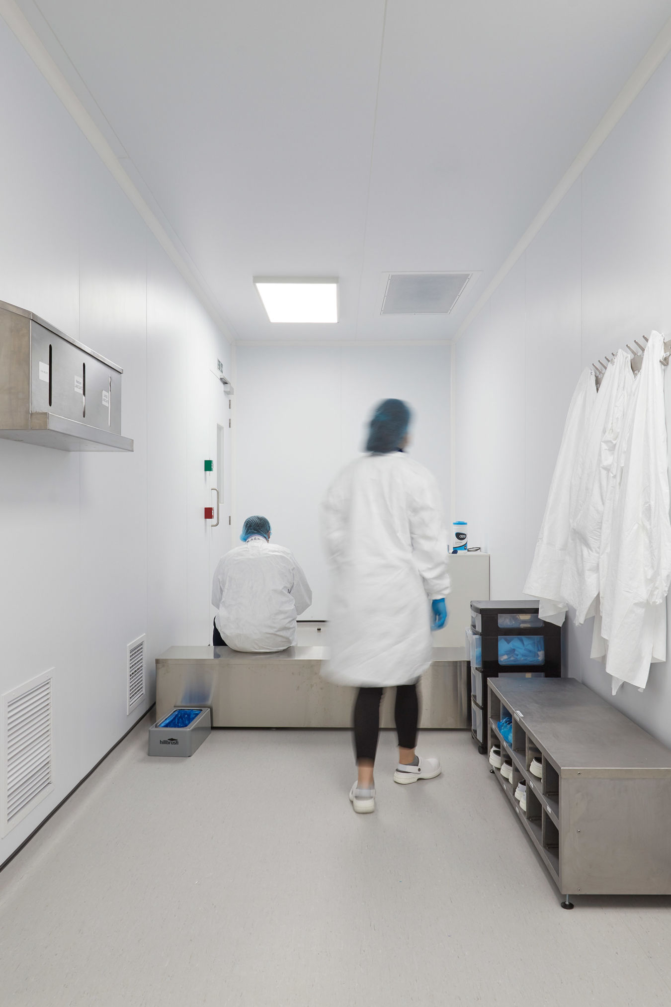 Cleanroom corridor with white walls and bright lighting, featuring lab personnel in protective clothing, wall-mounted lab coats, and a stainless steel bench and storage area.
