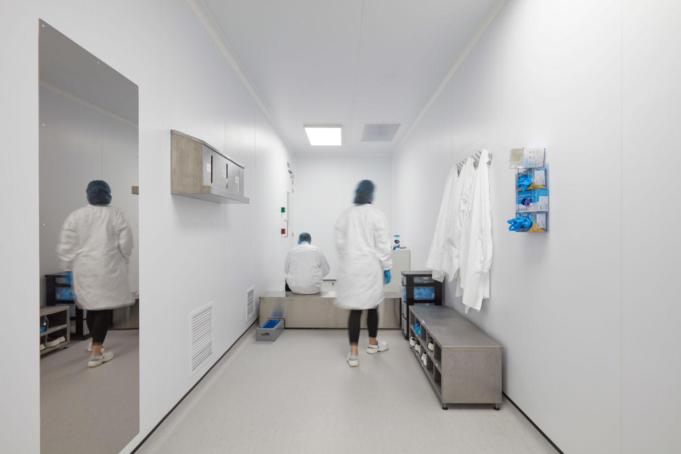 Cleanroom corridor with white walls and bright lighting, featuring lab personnel in protective clothing, wall-mounted lab coats, and a stainless steel bench and storage area.