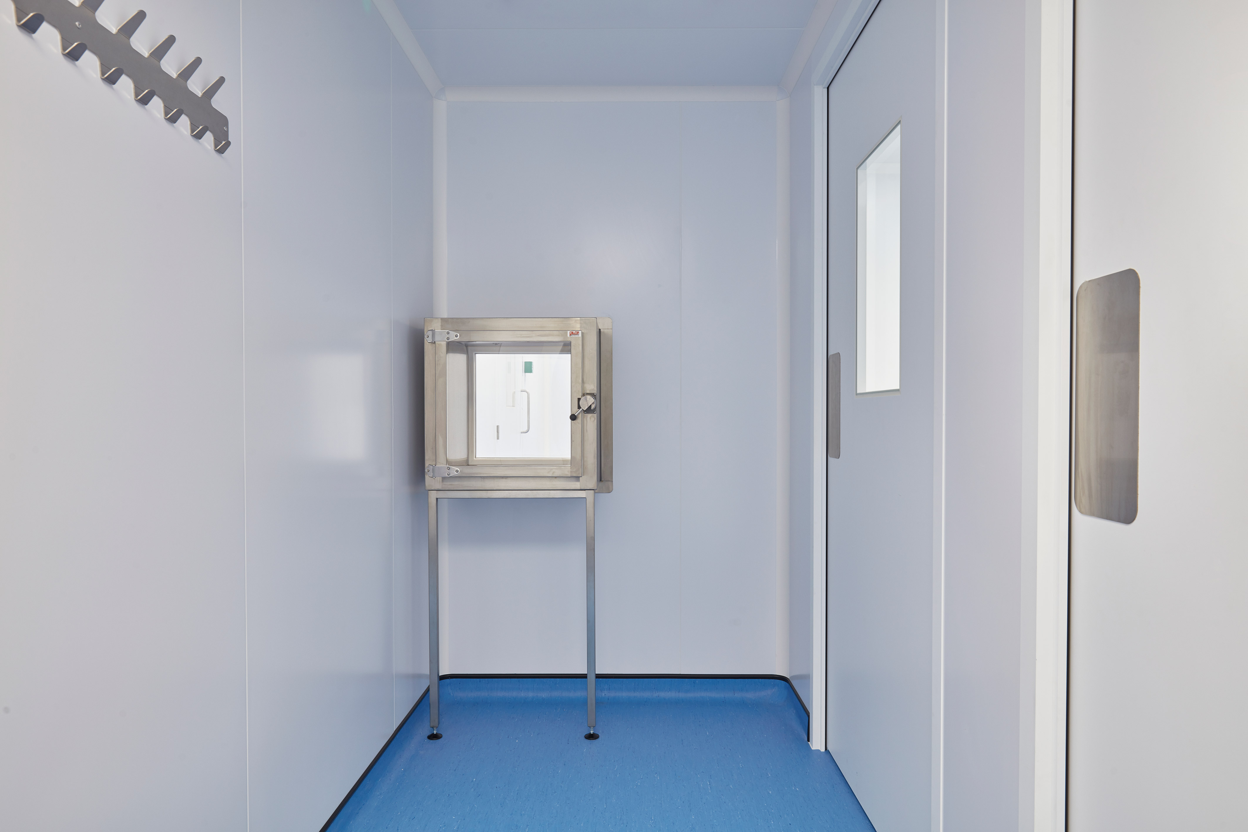 Cleanroom entry area with light blue walls, a pass-through chamber mounted on metal legs, and a sliding door with a small observation window.