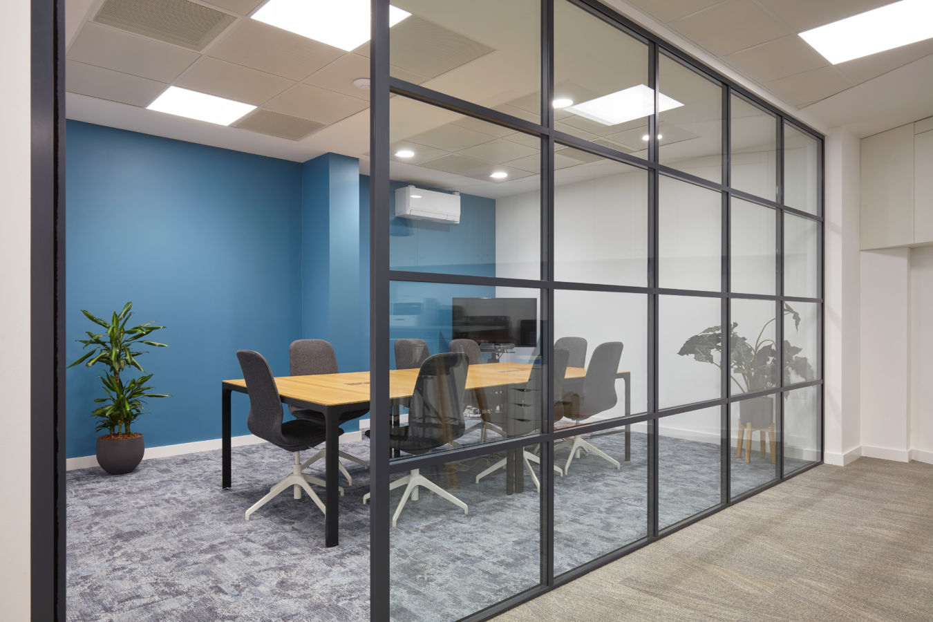 Modern office meeting room with a glass wall, featuring a wooden table, eight gray chairs, a blue accent wall, potted plants, and soft lighting.