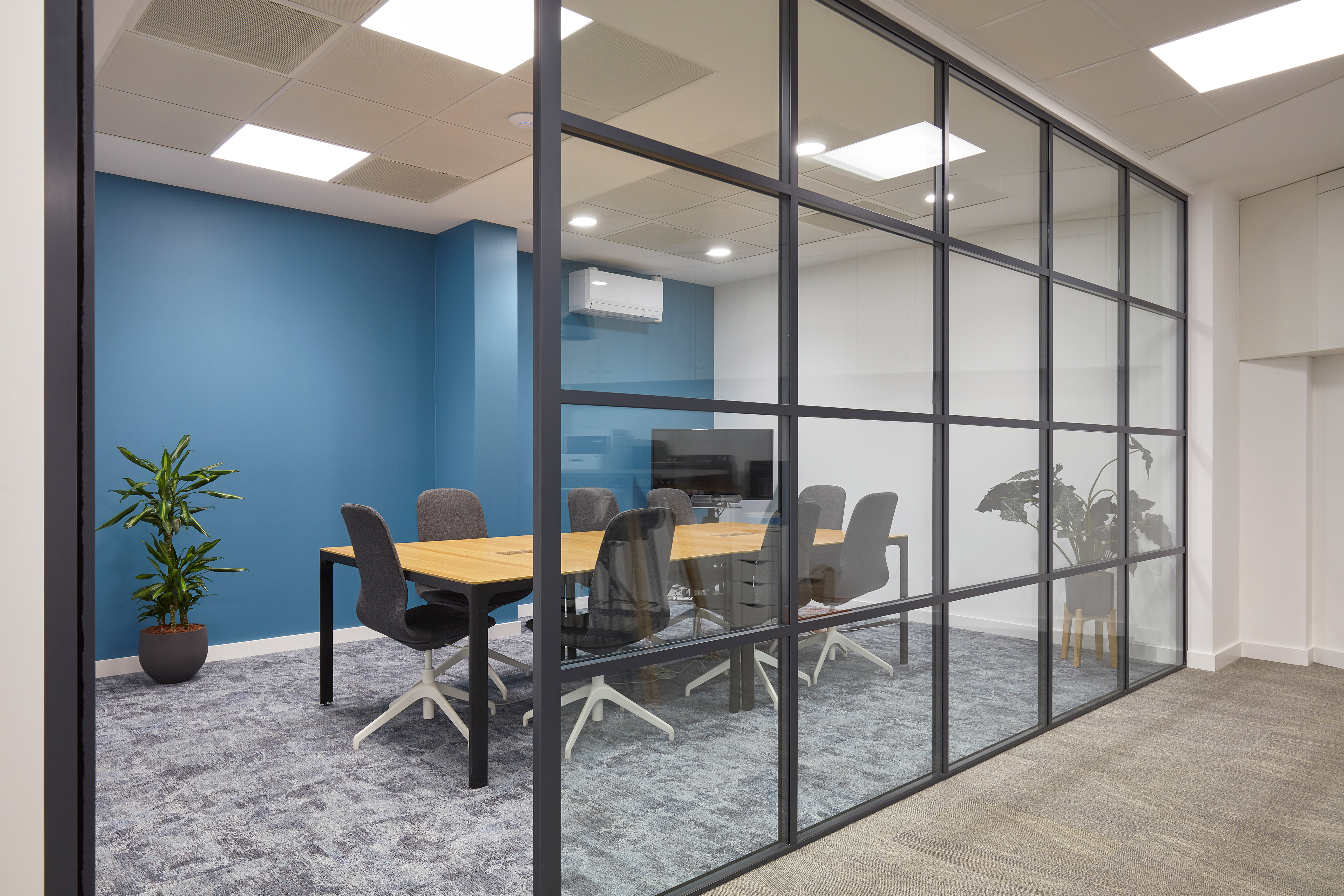 Modern office meeting room with a glass wall, featuring a wooden table, eight gray chairs, a blue accent wall, potted plants, and soft lighting.