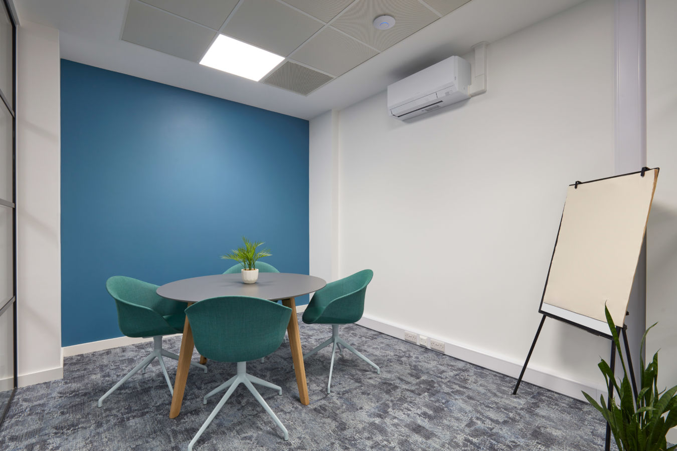 Small meeting room with a blue accent wall, round gray table, and three teal chairs. A blank easel stands nearby, and a small plant decorates the table