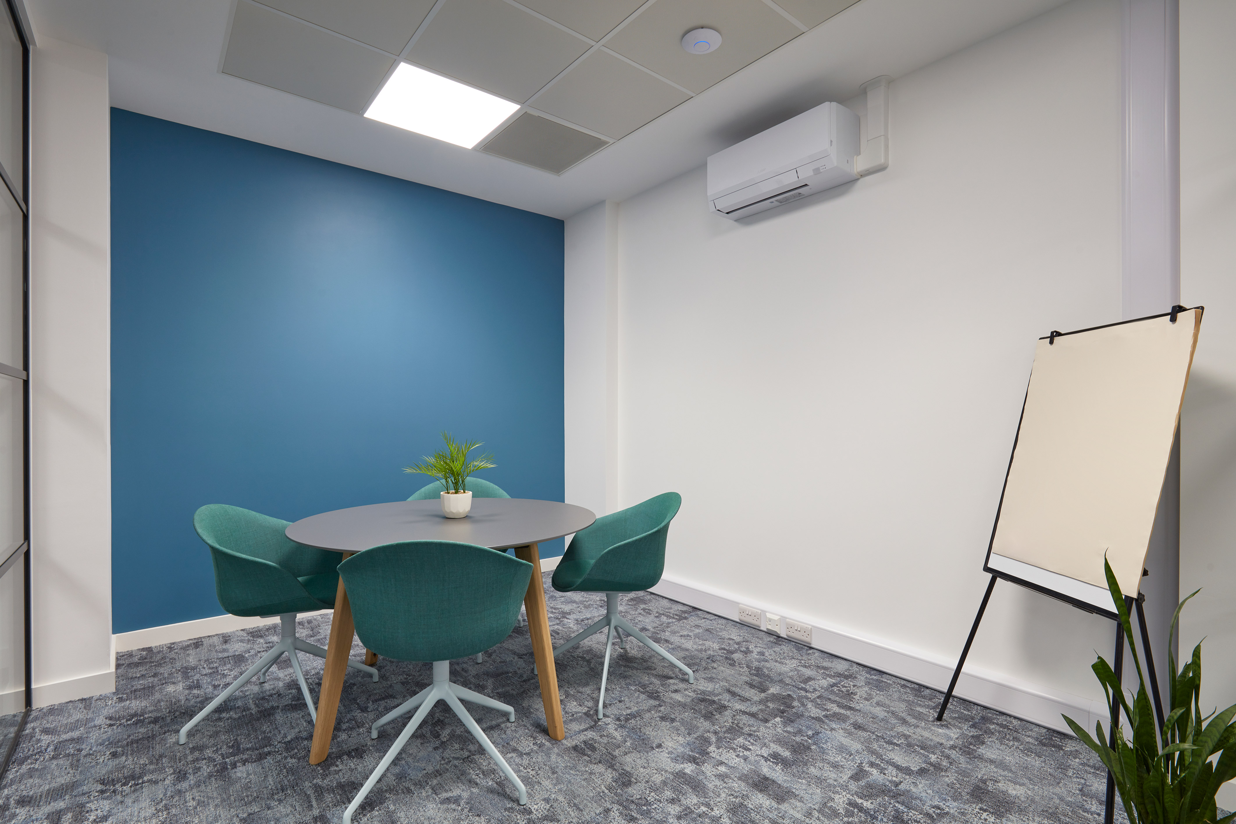 Small meeting room with a blue accent wall, round gray table, and three teal chairs. A blank easel stands nearby, and a small plant decorates the table