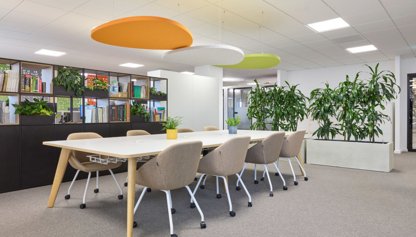 Modern office space with a large table and beige chairs. Ceiling panels in orange, white, and green add color. Bookshelves and plants create a fresh atmosphere