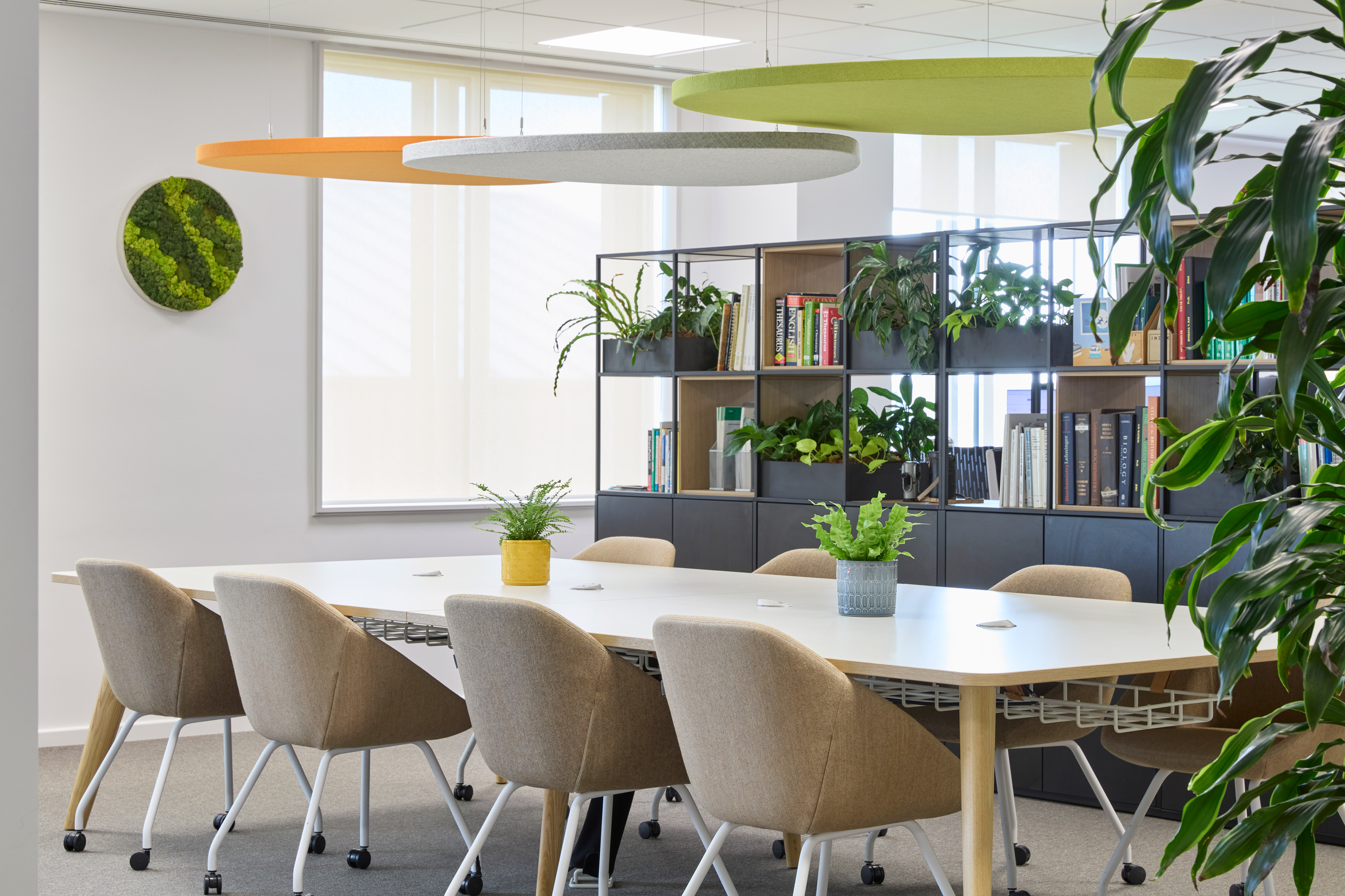 Modern office meeting room with a long white table, beige chairs, and plants. Shelves with books and more greenery create a fresh, inviting atmosphere