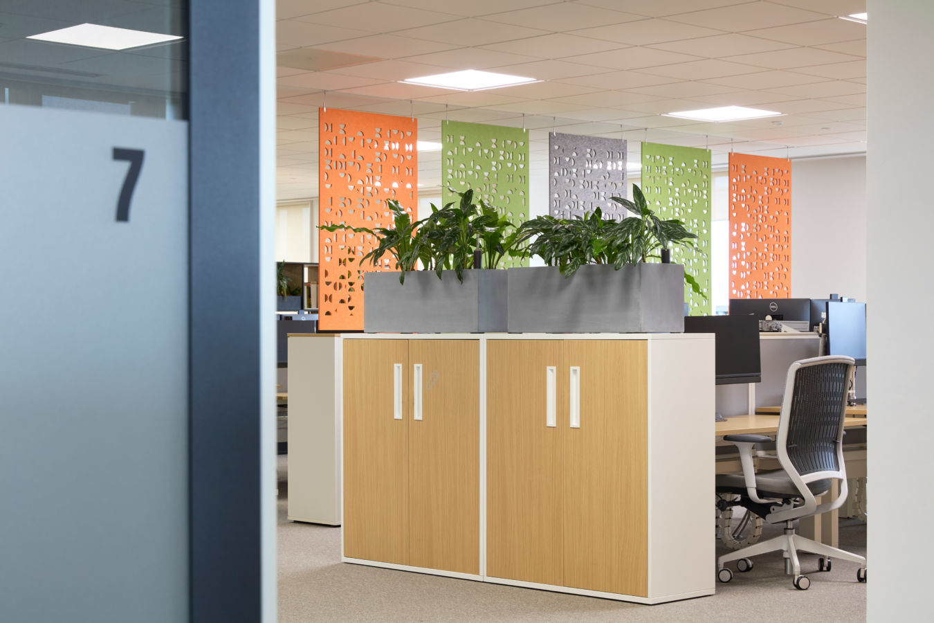 Office scene features plants on cabinets, surrounded by colorful panels and workstations with chairs and computers. Door labeled with the number "7" within sleek, modern workspace.