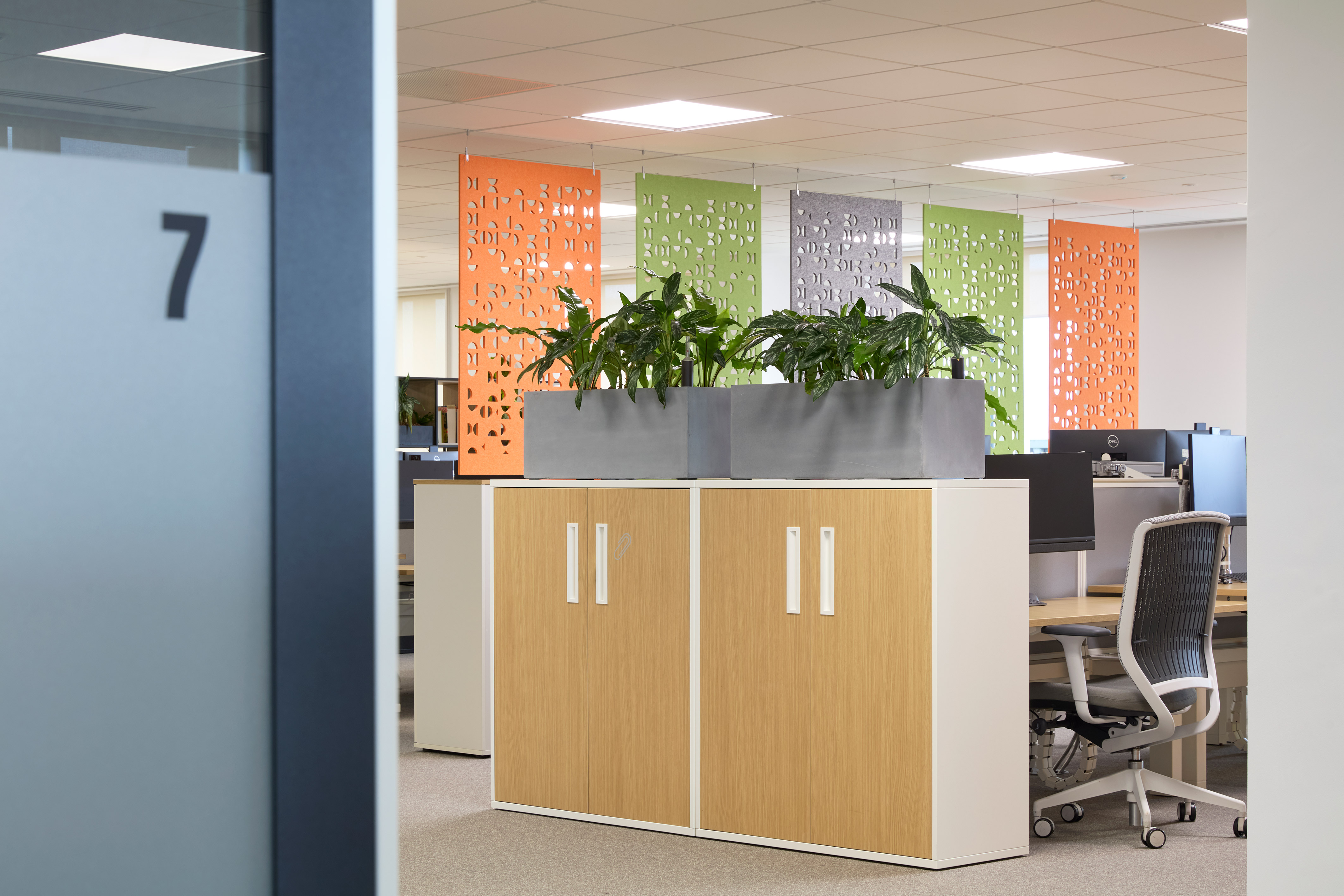 Office scene features plants on cabinets, surrounded by colorful panels and workstations with chairs and computers. Door labeled with the number "7" within sleek, modern workspace.