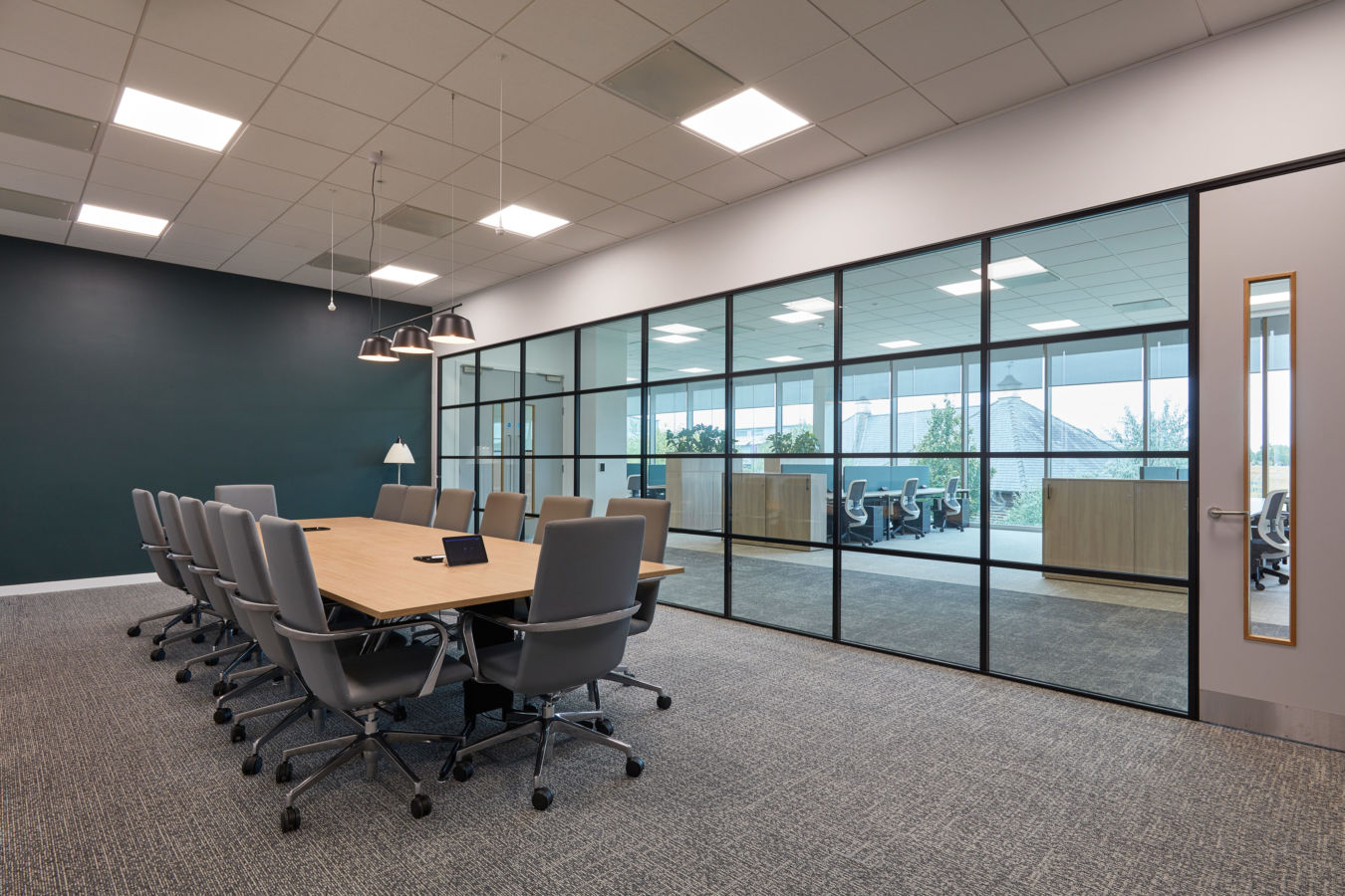 A large wooden conference table surrounded by gray chairs is set in a modern meeting room with carpeted floors, pendant lights, and glass walls overlooking an office area.