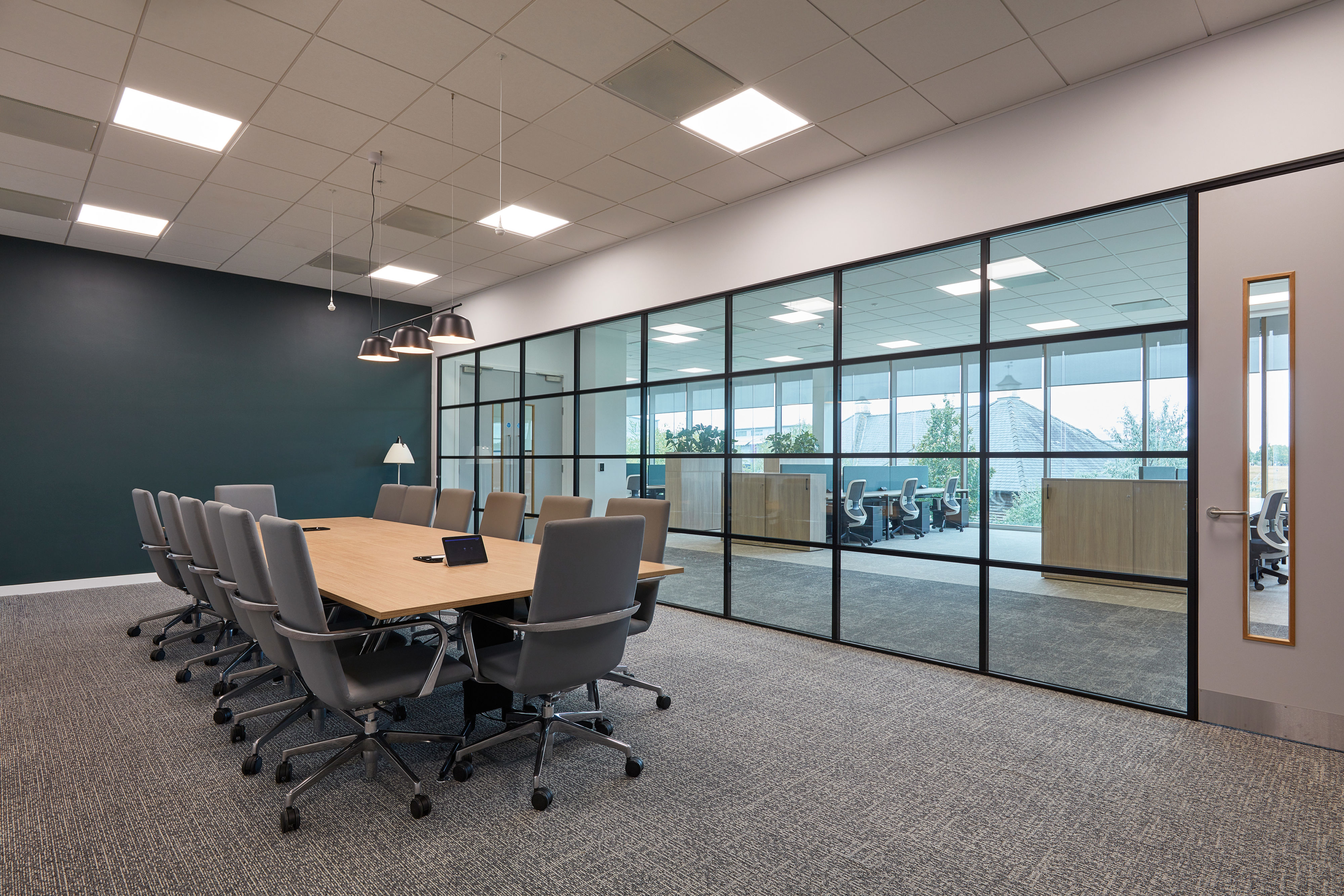 A large wooden conference table surrounded by gray chairs is set in a modern meeting room with carpeted floors, pendant lights, and glass walls overlooking an office area.