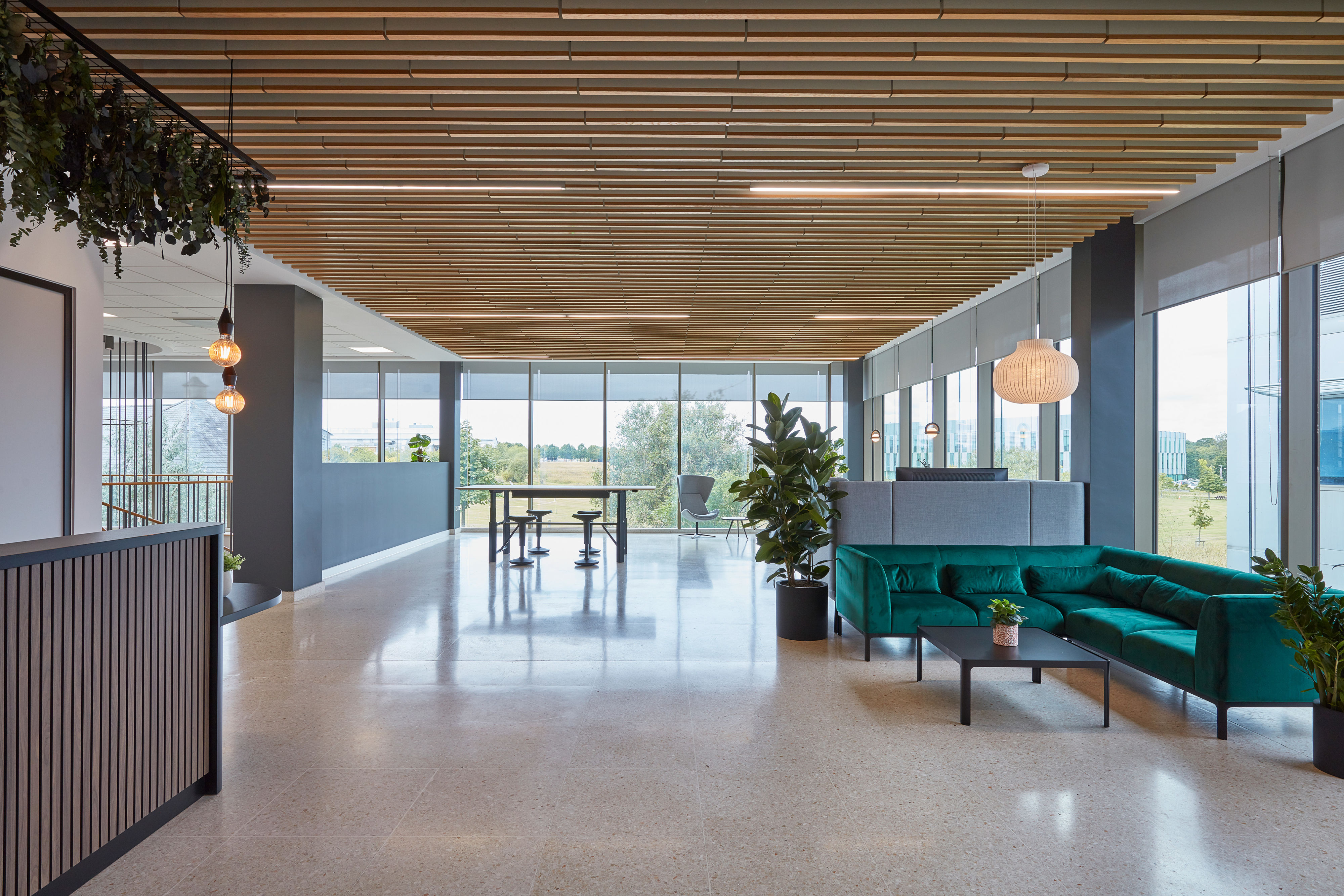 Green velvet sofa sits idle, surrounded by potted plants and wooden furniture, in a modern office space with large windows and ceiling lights.