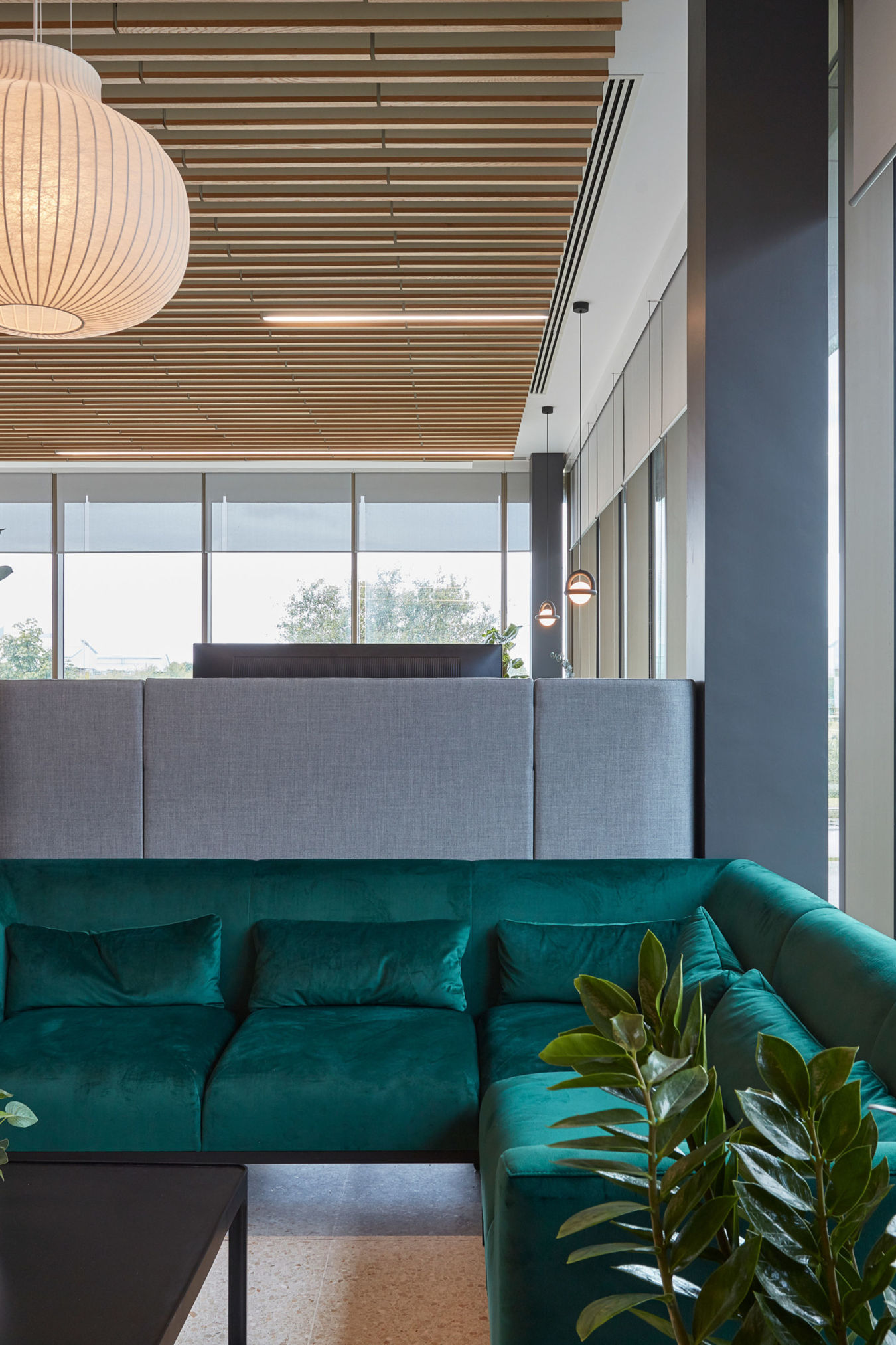 A modern office lounge features a teal sectional sofa surrounded by large green plants, beneath a wooden ceiling with a spherical pendant light, next to large windows revealing an outdoor view.