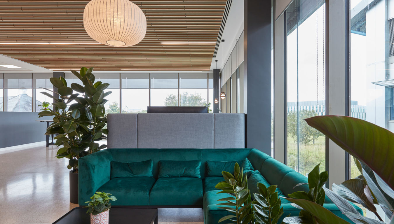 A modern office lounge features a teal sectional sofa surrounded by large green plants, beneath a wooden ceiling with a spherical pendant light, next to large windows revealing an outdoor view.