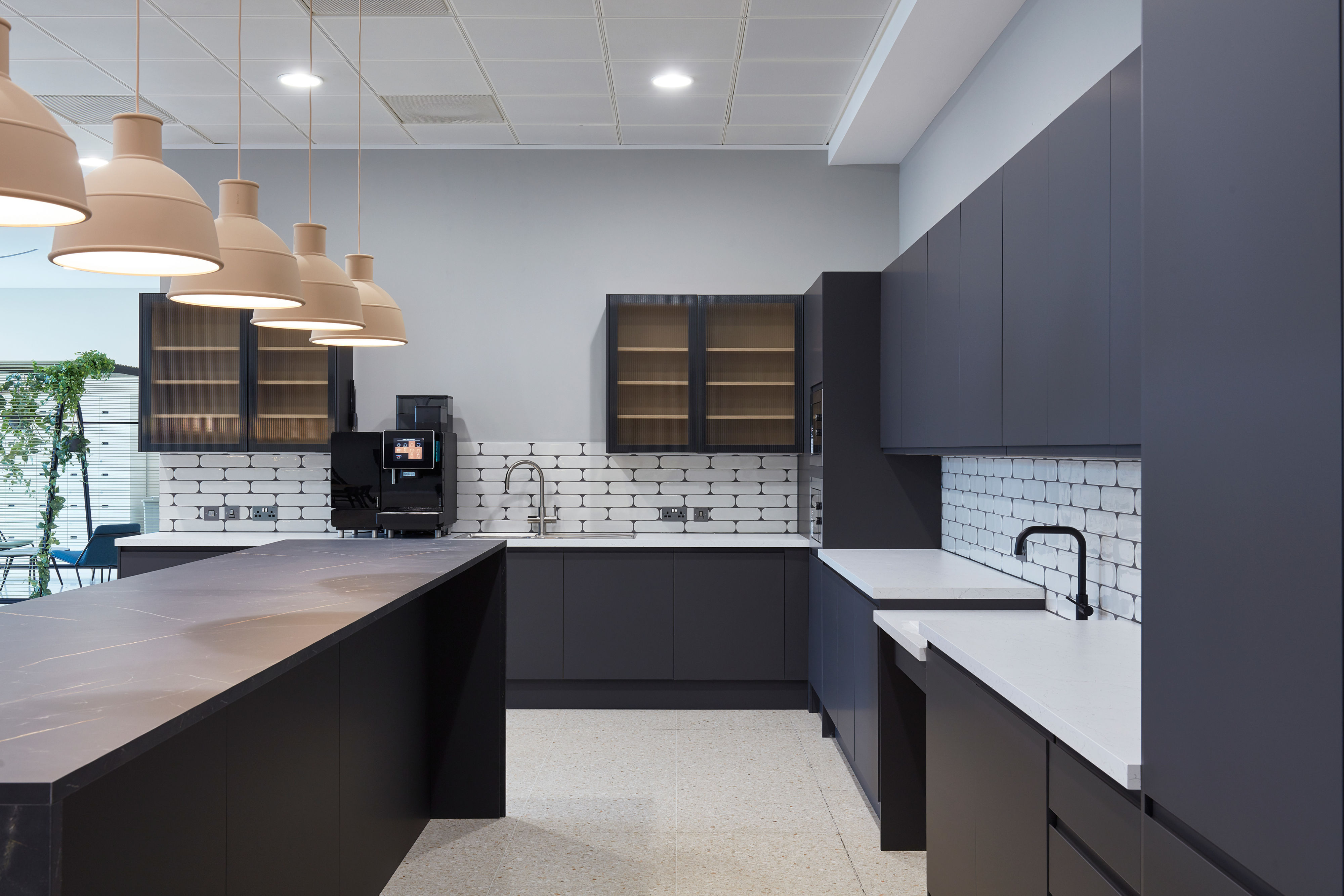 Modern kitchen features sleek black cabinetry, a large island with beige pendant lights, and a coffee machine on a white-tiled backsplash, providing a contemporary, minimalist aesthetic.