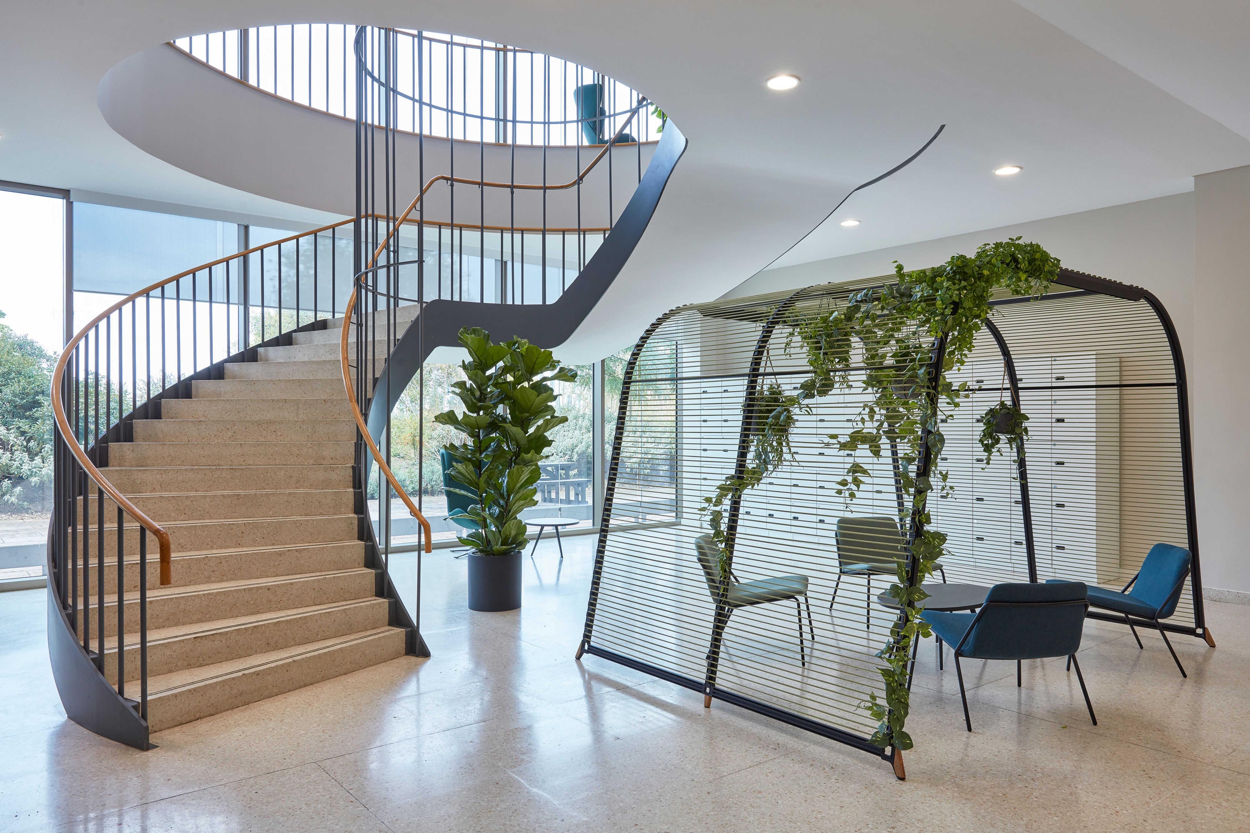 A spiral staircase with wooden railings ascends next to a modern seating area enclosed by a slatted structure adorned with green plants, in a well-lit atrium with large windows.
