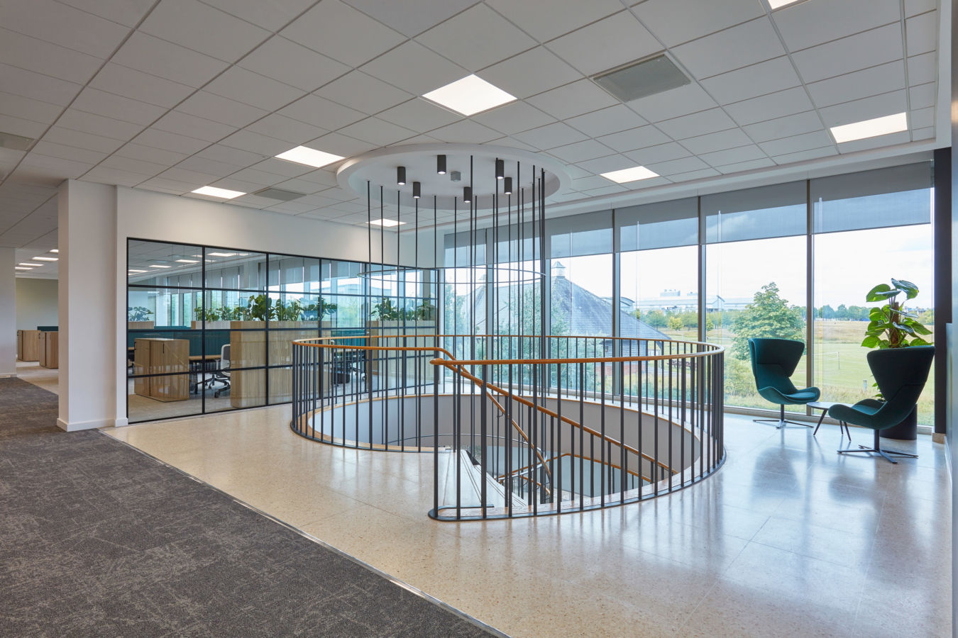 Open spiral staircase with wooden handrails and metal railings descends in a modern office space, featuring glass walls, green chairs, a potted plant, and expansive windows overlooking greenery.