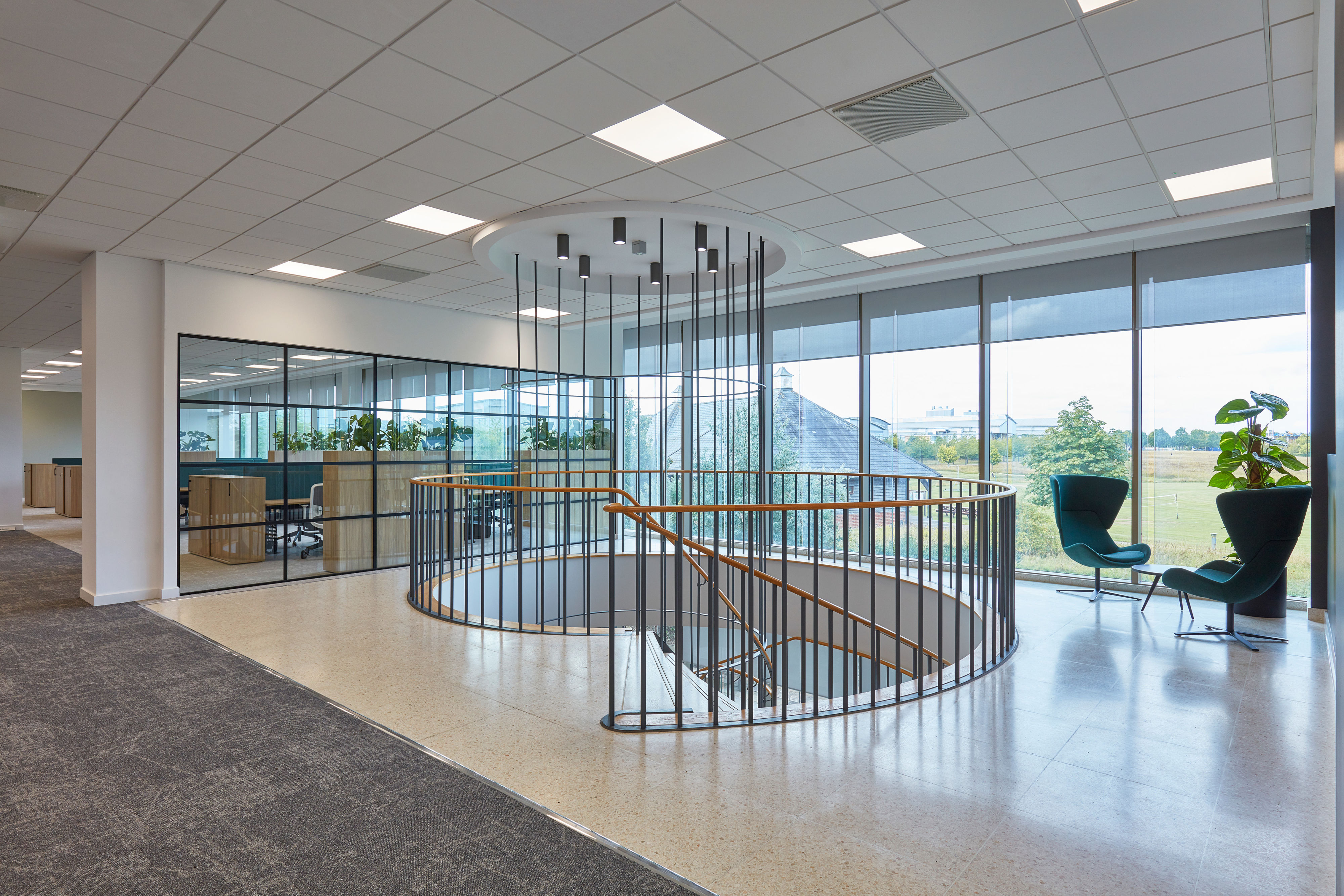 Open spiral staircase with wooden handrails and metal railings descends in a modern office space, featuring glass walls, green chairs, a potted plant, and expansive windows overlooking greenery.