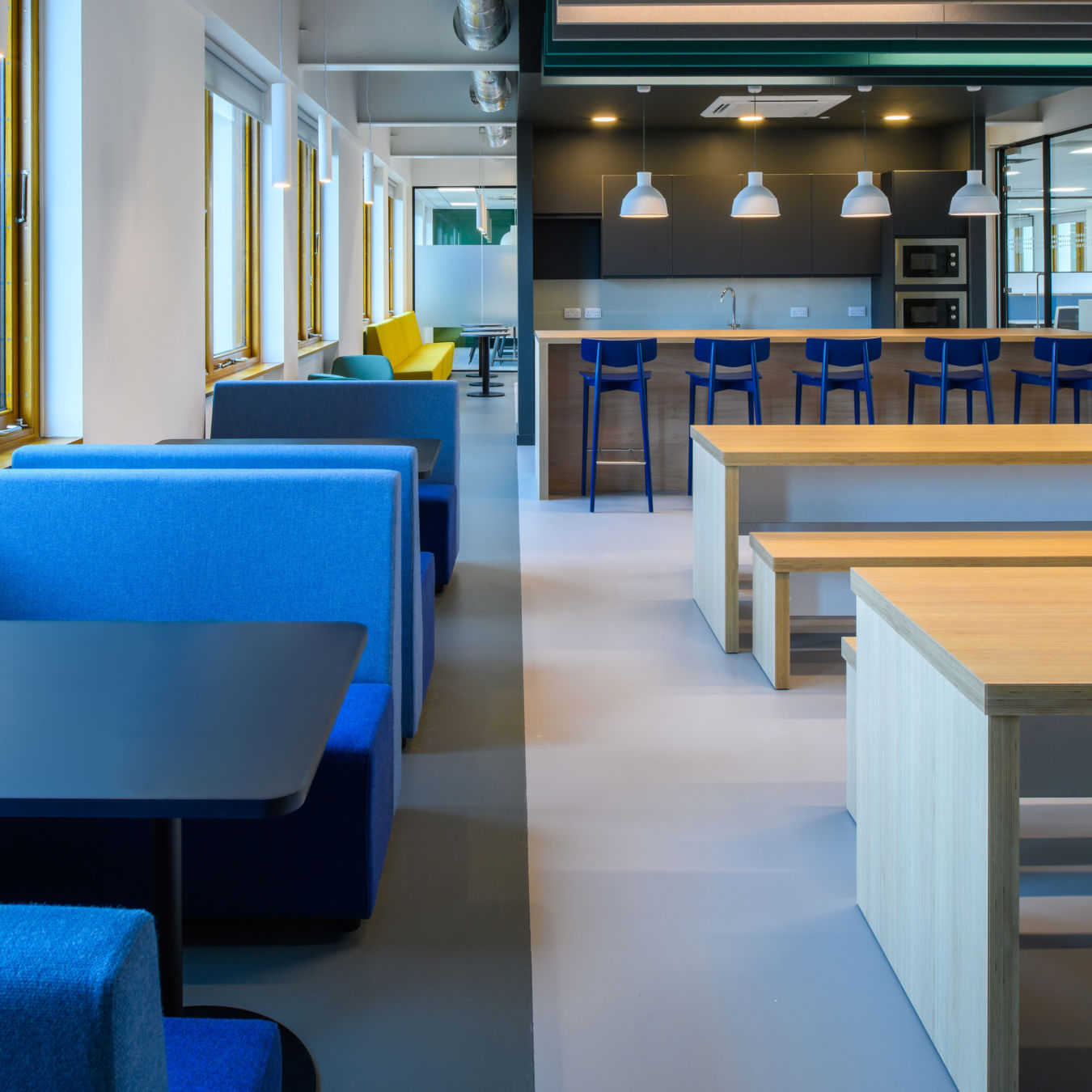 Modern office kitchen with blue and yellow seating, wooden tables, and bar stools. Bright lighting, large windows, and a calm, inviting atmosphere