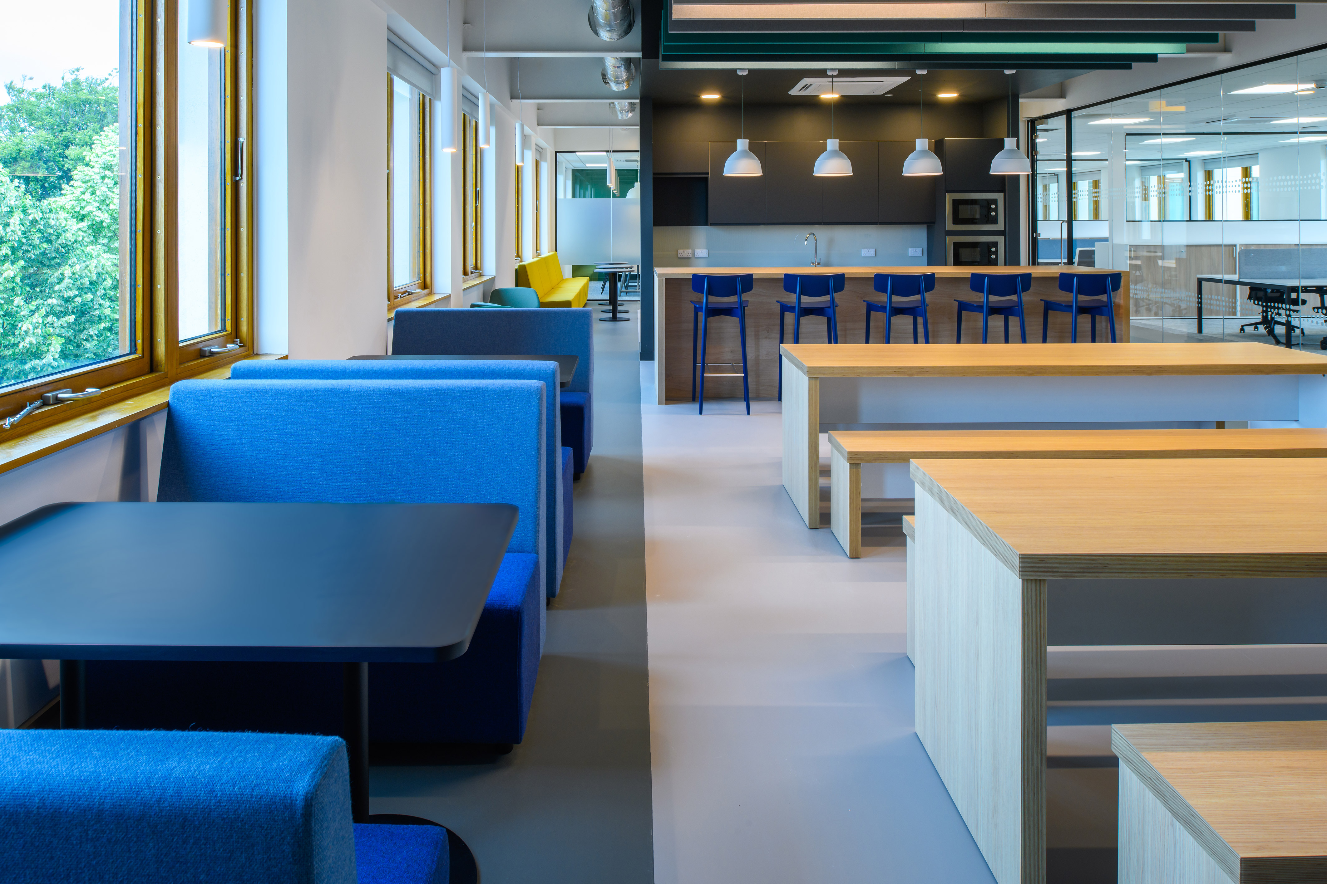 Modern office kitchen with blue and yellow seating, wooden tables, and bar stools. Bright lighting, large windows, and a calm, inviting atmosphere