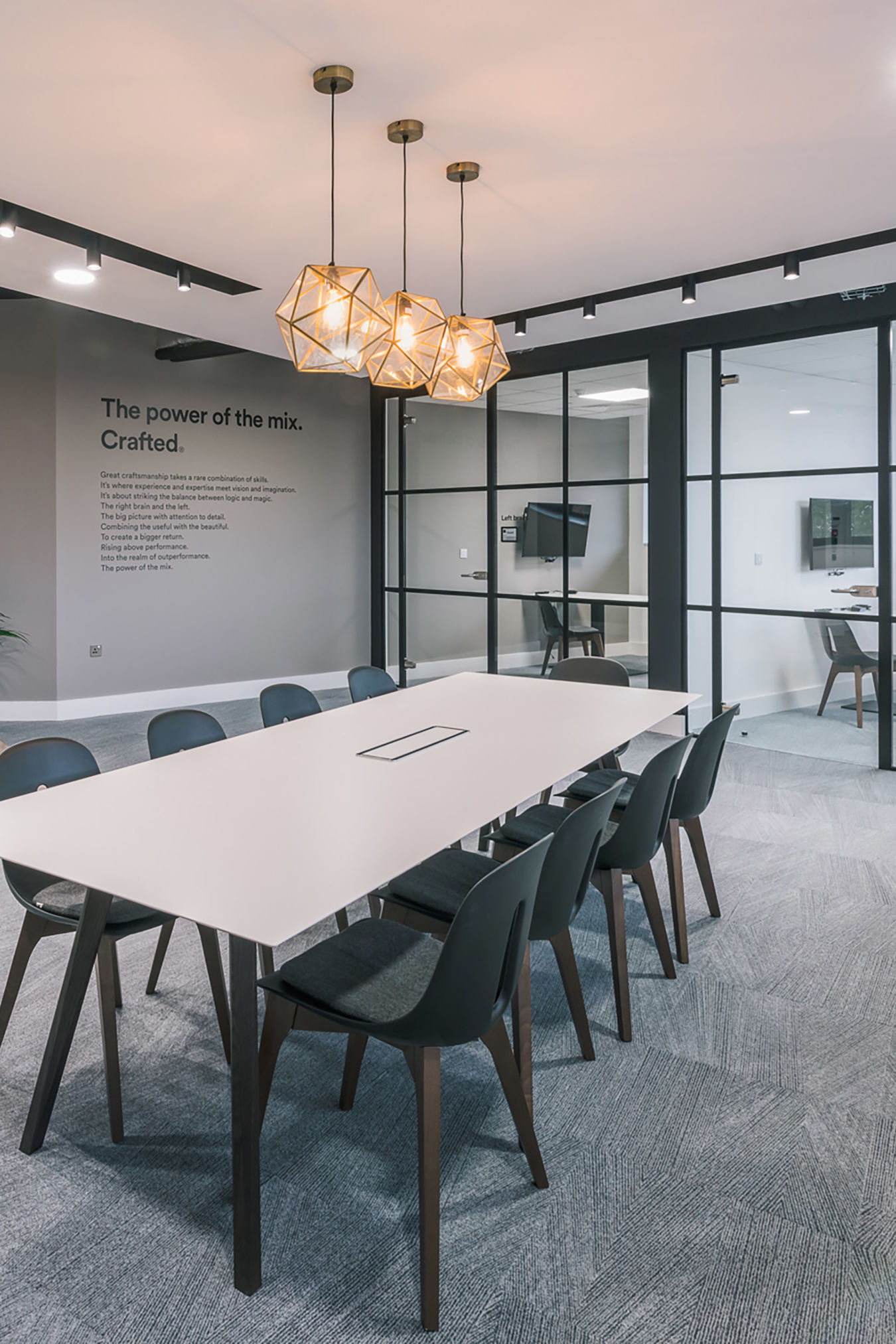 Modern office with a sleek conference room featuring a long white table and black chairs. Glass walls reveal an adjacent workspace. Elegant pendant lights and greenery add warmth.