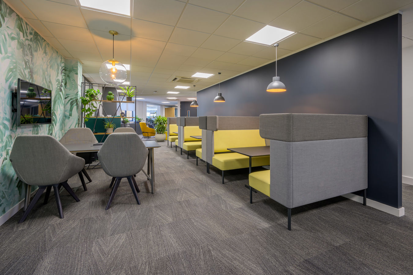 Modern office lounge with gray and yellow booth seating, leafy wallpaper, and pendant lighting. Grey chairs and a wall-mounted TV add sophistication.
