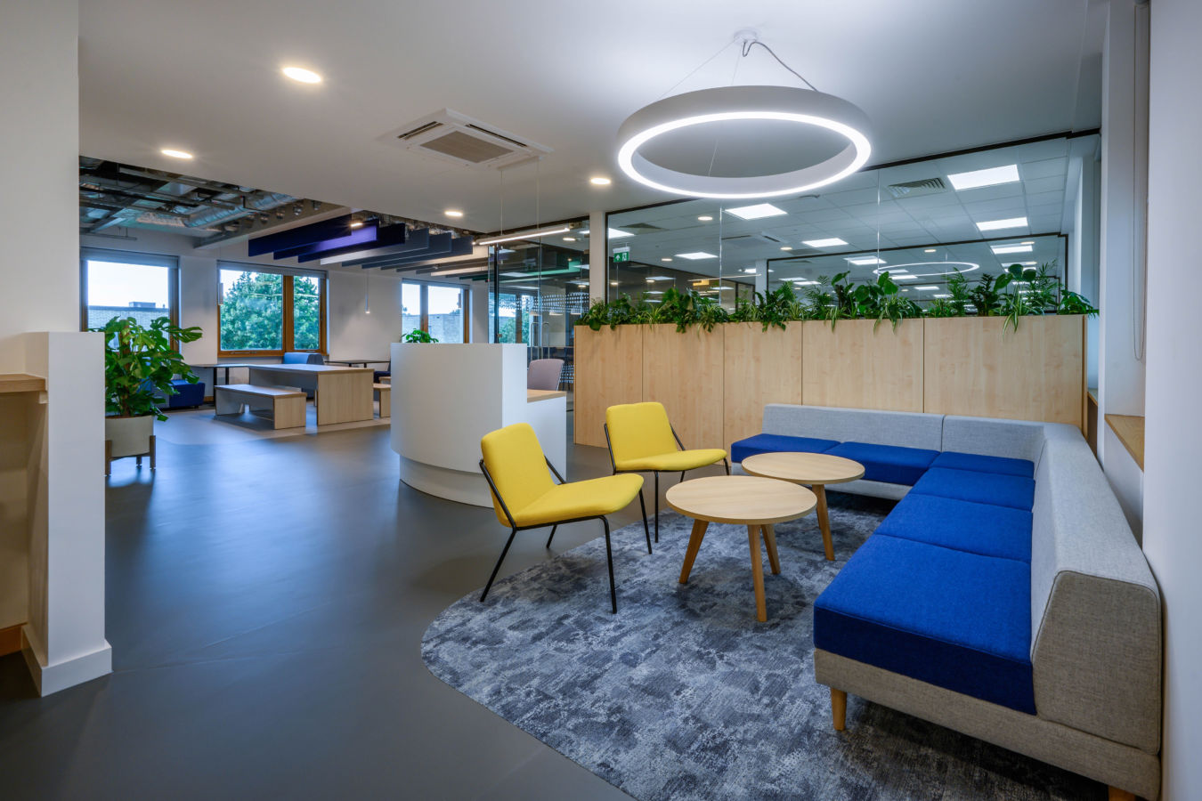 Yellow chairs and round wooden tables are arranged on a textured rug in a modern office lounge. A circular light hangs above, with green plants lining a partition wall.