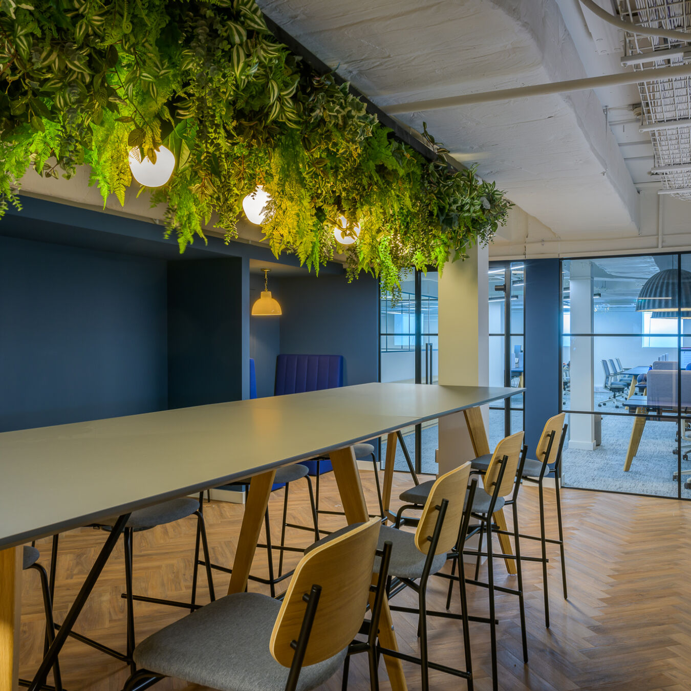Modern office interior with a long table surrounded by wooden chairs. Green plants and round lights hang above, creating a fresh, inviting atmosphere