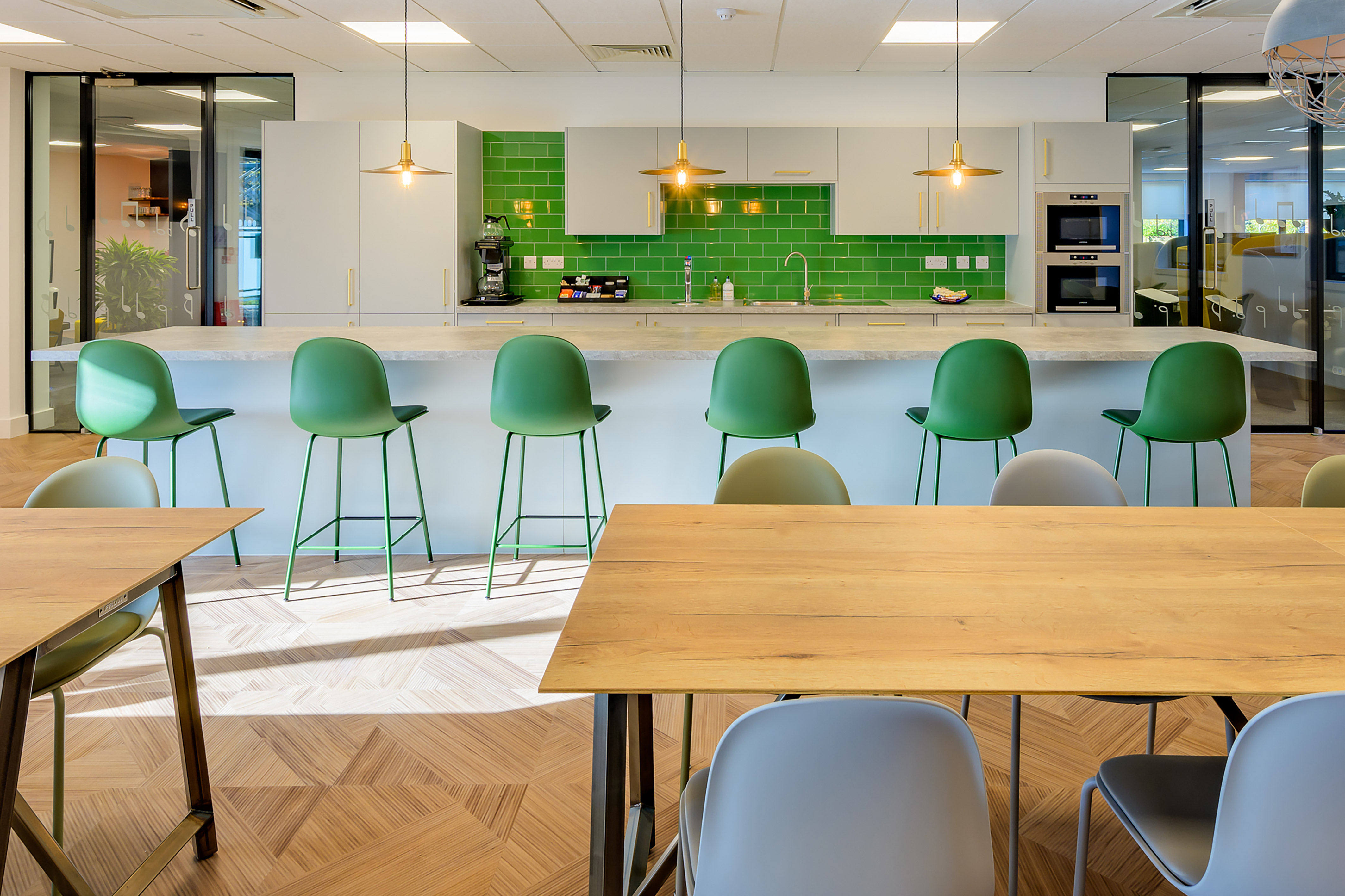 Modern kitchen with green chairs lined up at a white countertop; green-tiled backsplash, white cabinets, and pendant lights create a stylish office break area setting.