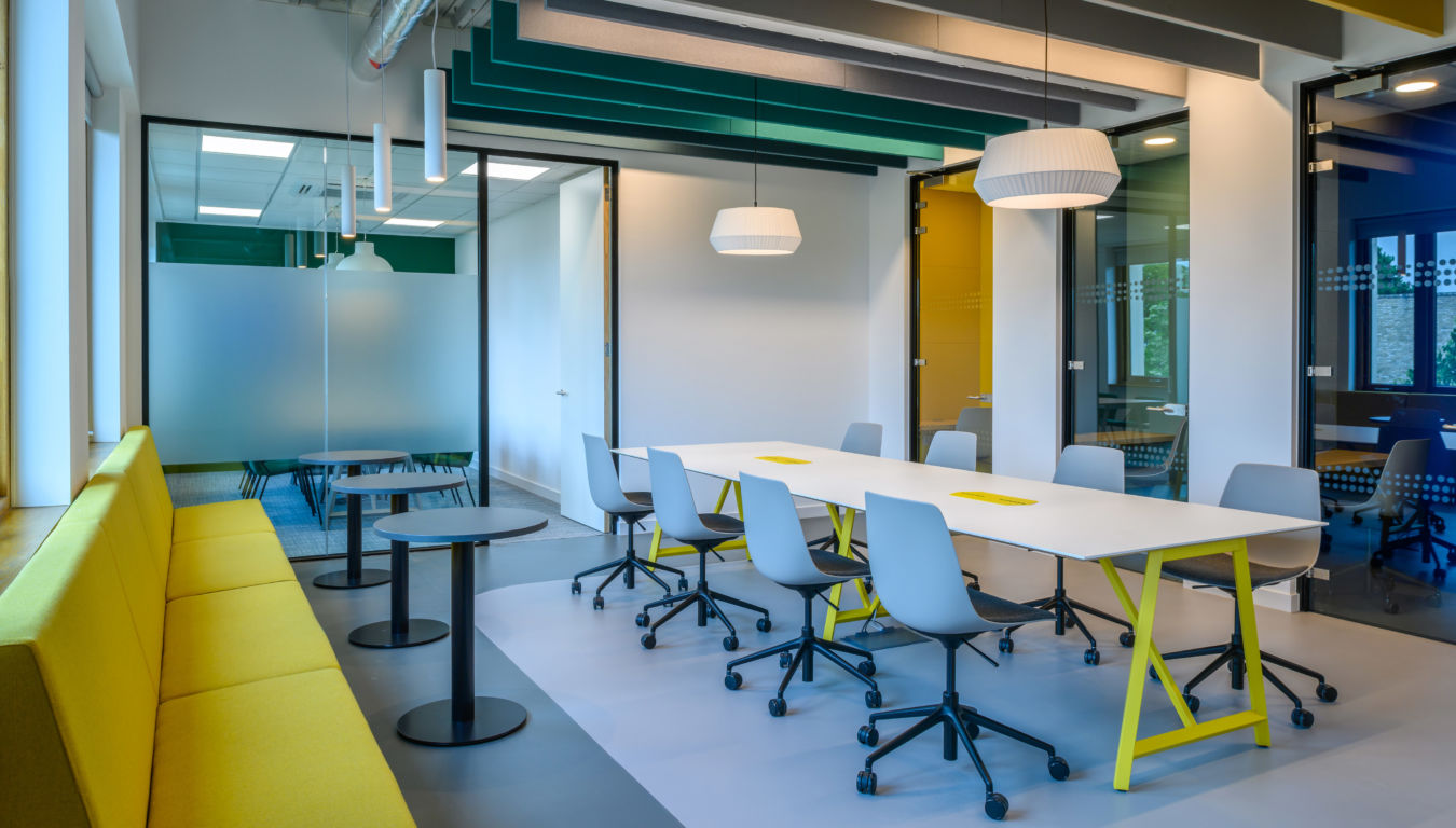 Meeting room with a long white table and gray chairs arranged neatly. Yellow bench seating lines one wall, pendant lights hang overhead, and frosted glass partitions divide the space.