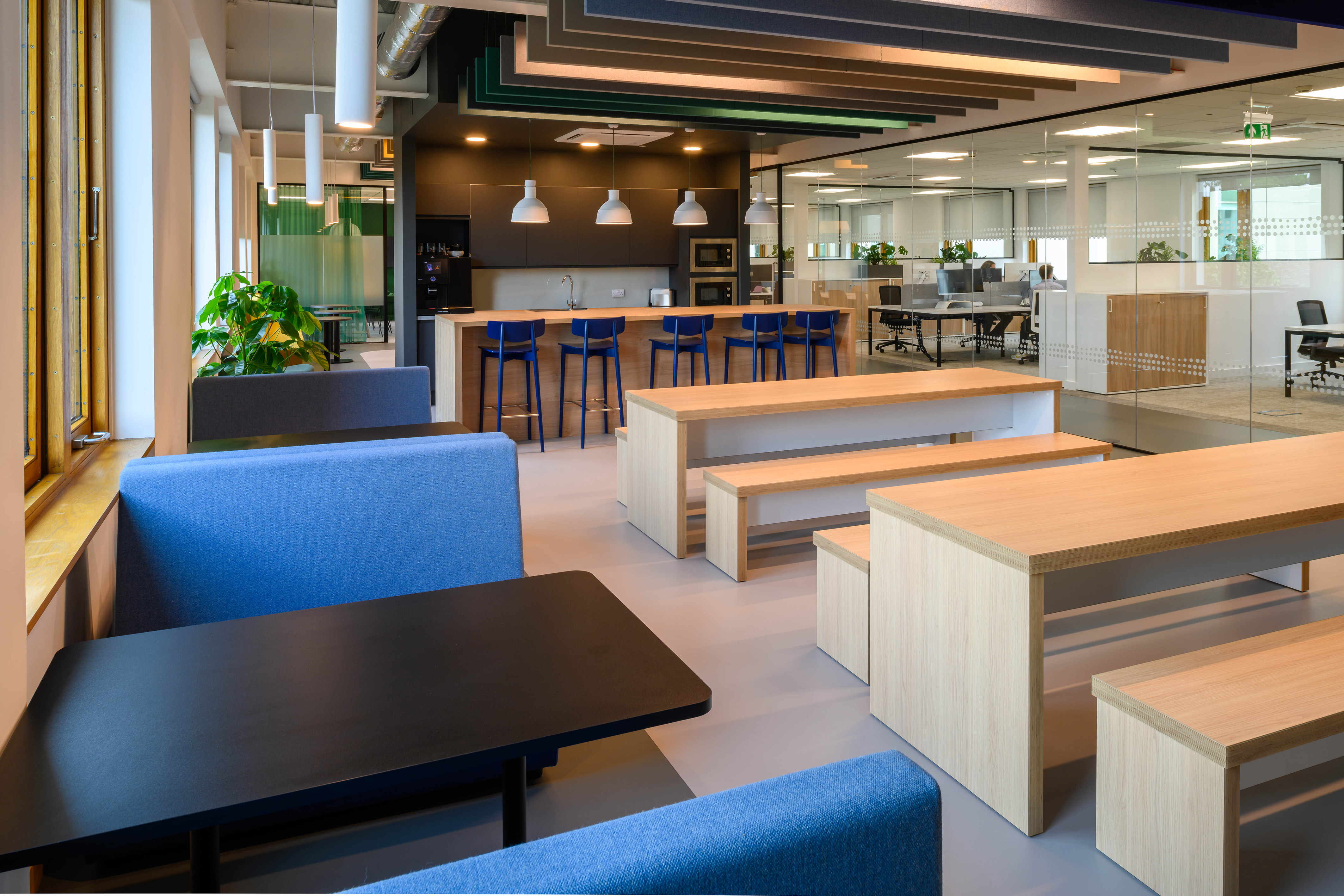 Modern office lounge with blue sofas, wooden tables, bar seating, and pendant lights. Open layout with glass walls and greenery adds a relaxed vibe