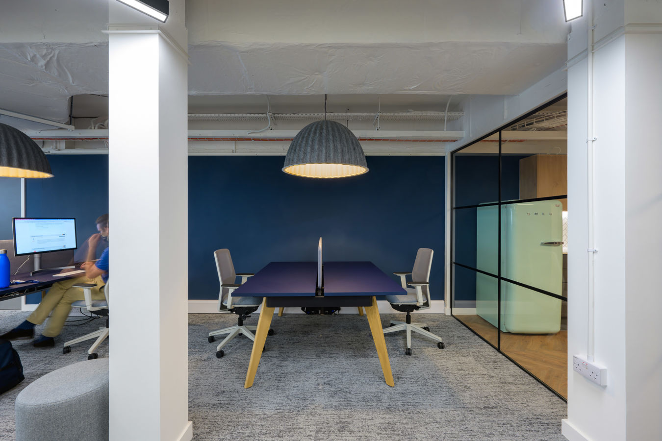 Office interior features a modern desk with two chairs and a central divider, illuminated by a pendant lamp. A person works at a computer with a blue water bottle nearby.