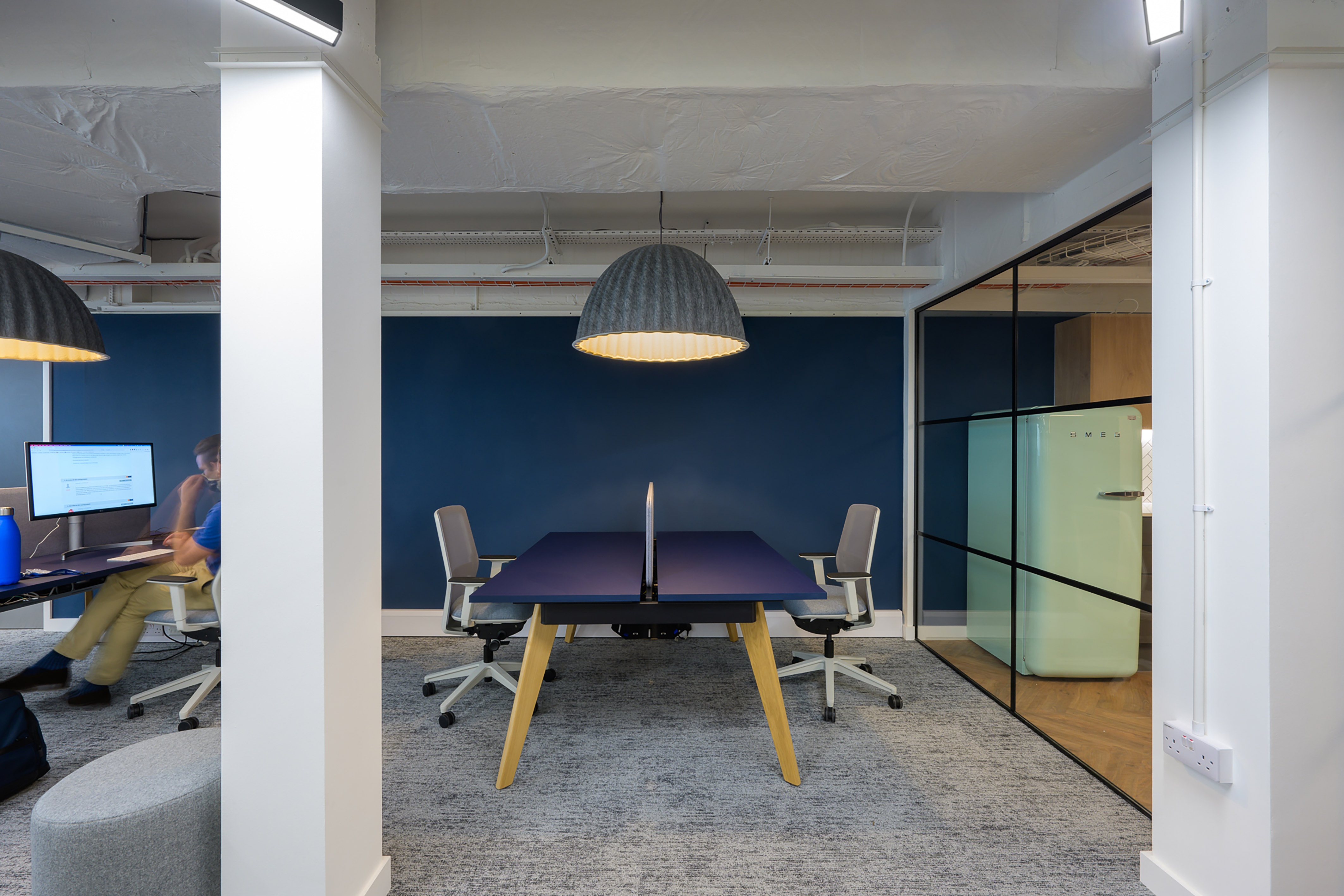 Office interior features a modern desk with two chairs and a central divider, illuminated by a pendant lamp. A person works at a computer with a blue water bottle nearby.