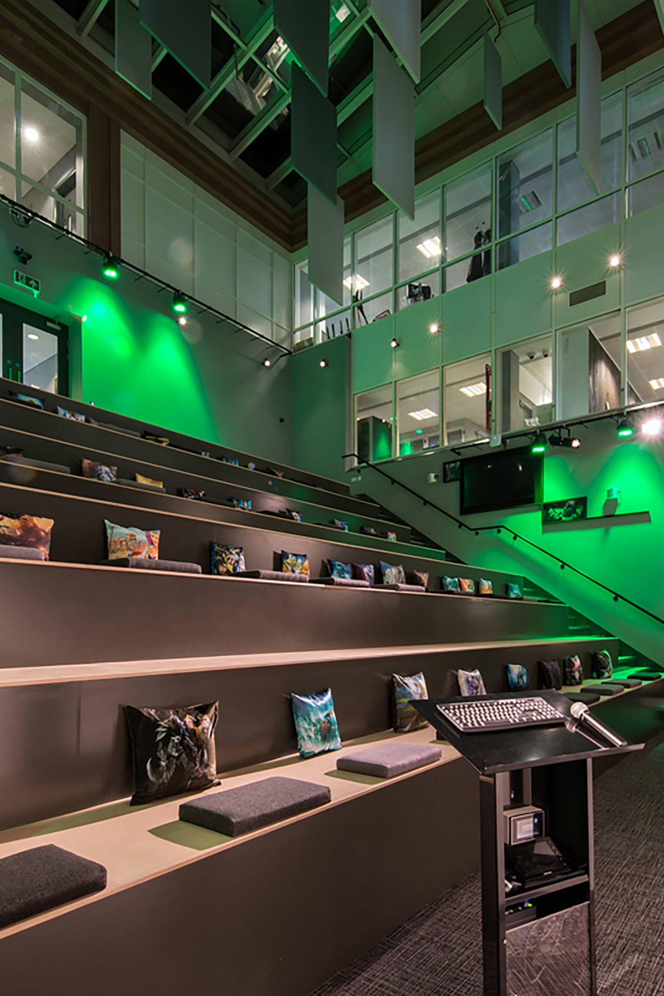 Auditorium with tiered wooden seats and colorful cushions, featuring a podium with a microphone and keyboard in the foreground, surrounded by green lighting and overlooking glass offices above.