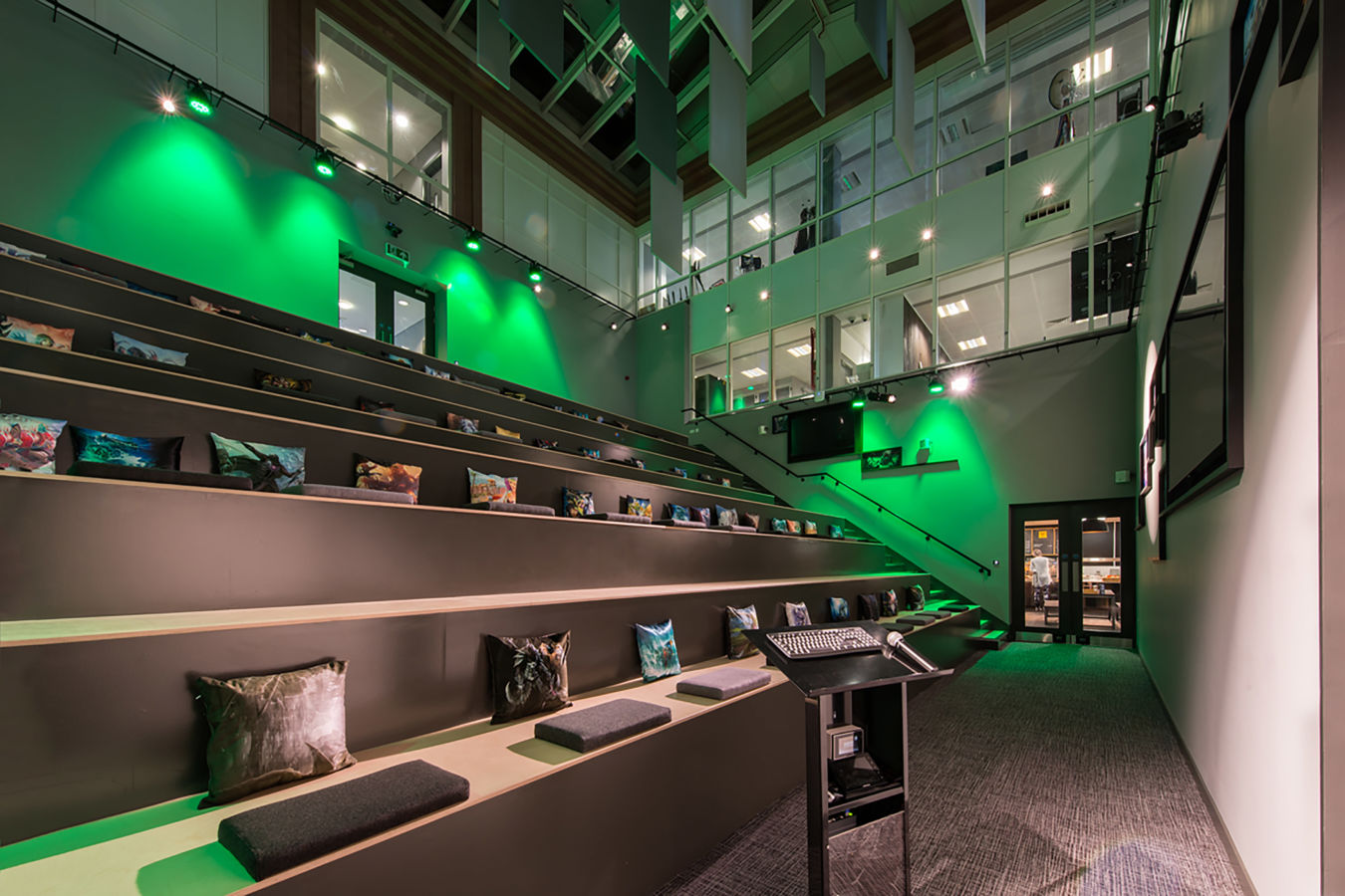 Auditorium with tiered wooden seats and colorful cushions, featuring a podium with a microphone and keyboard in the foreground, surrounded by green lighting and overlooking glass offices above.