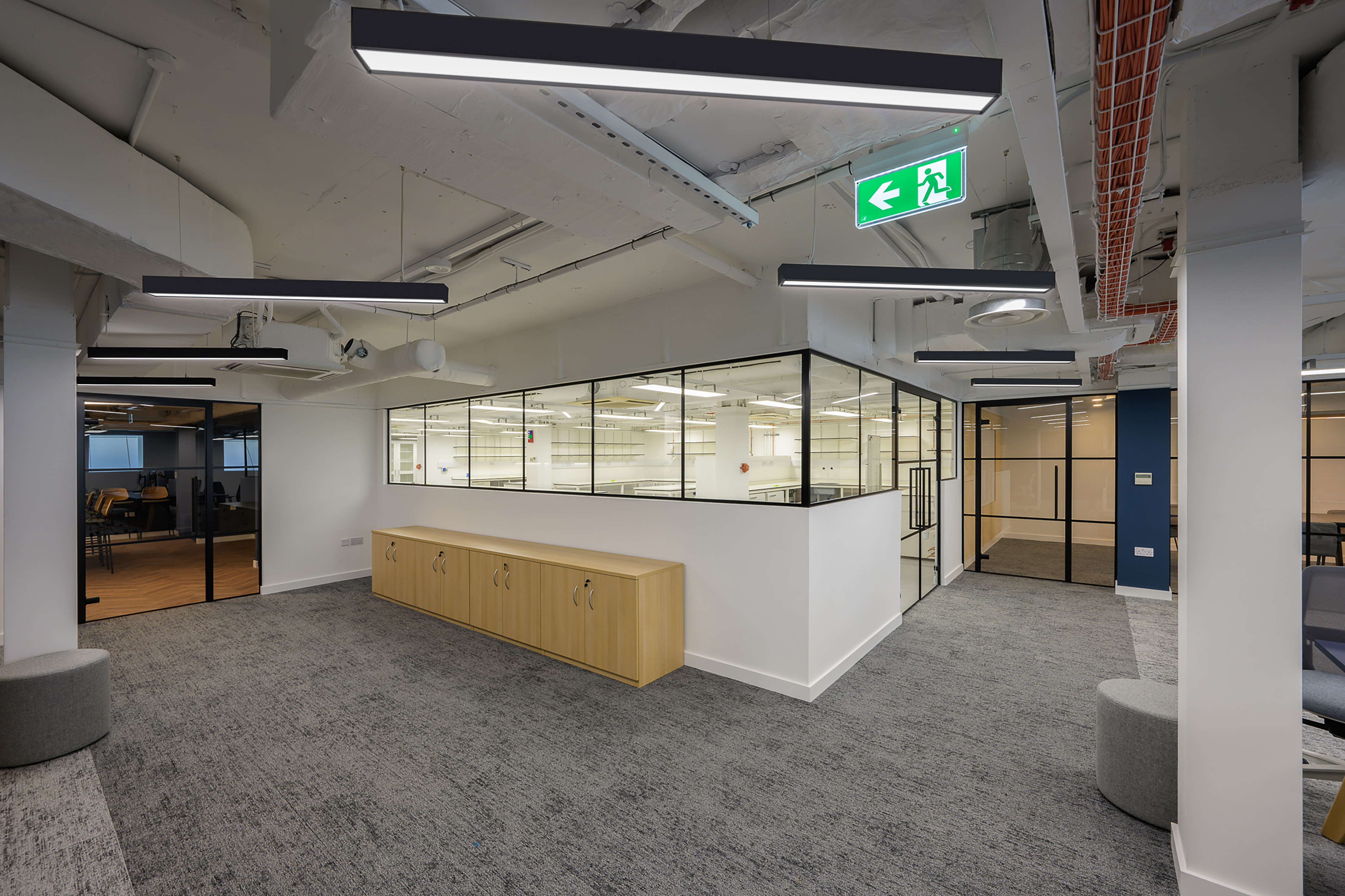 Modern office interior with gray carpet, white walls, and glass partitions. Wooden cabinets line a wall, and overhead lights create a sleek, professional feel