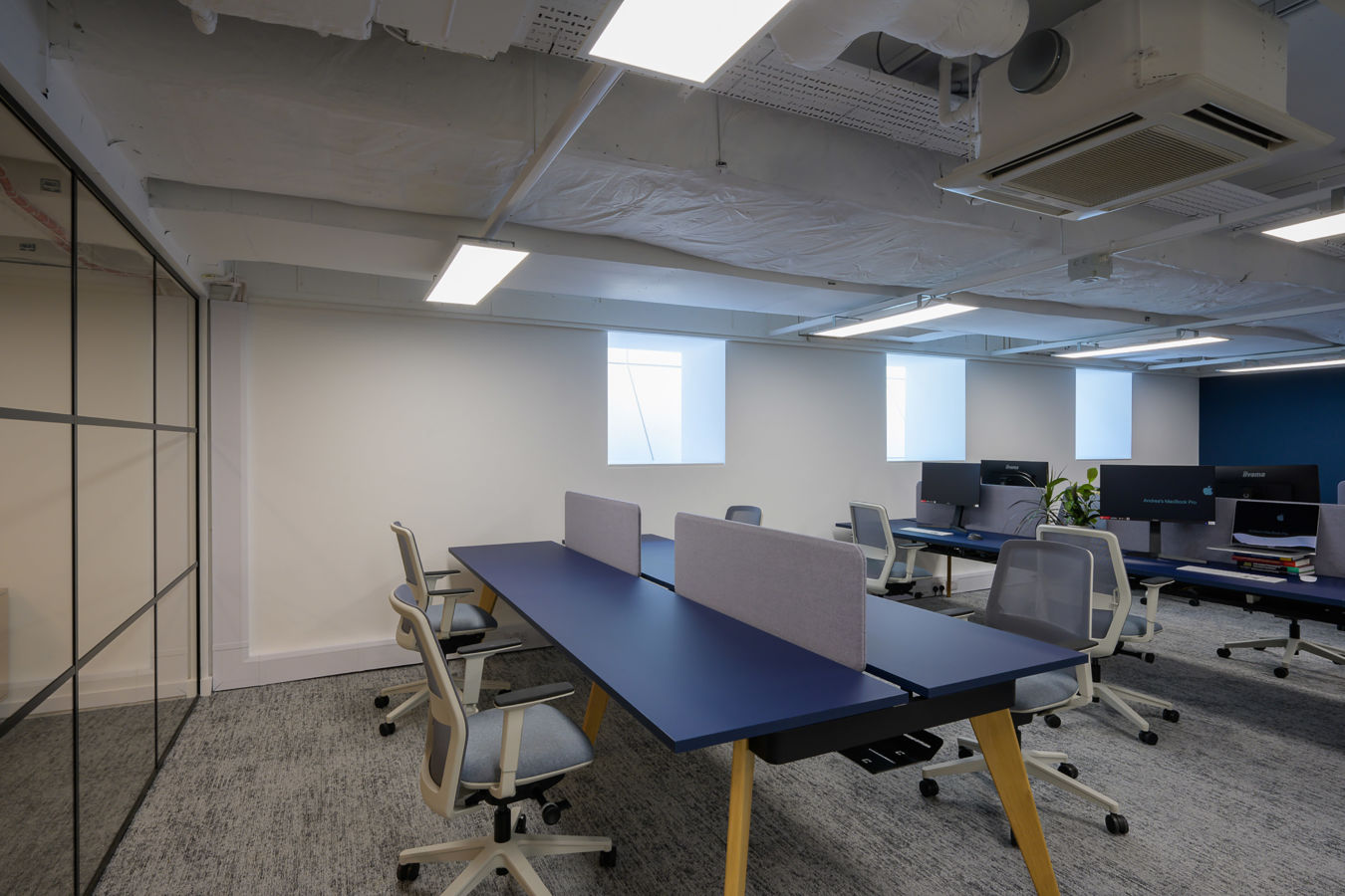 Modern office space with blue desks, gray chairs, and partitions. Overhead lighting and exposed ceiling add a minimalist feel. Neutral colors create a calm ambiance