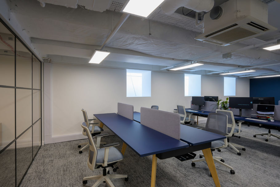 Modern office space with blue desks, gray chairs, and partitions. Overhead lighting and exposed ceiling add a minimalist feel. Neutral colors create a calm ambiance