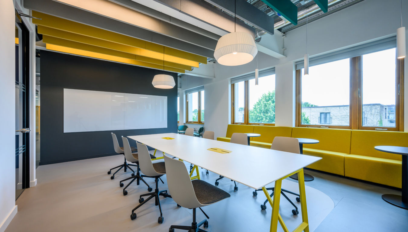 Modern office space with a long white table and gray chairs on wheels. Yellow accents include ceiling beams and a cushioned bench. Large windows provide natural light