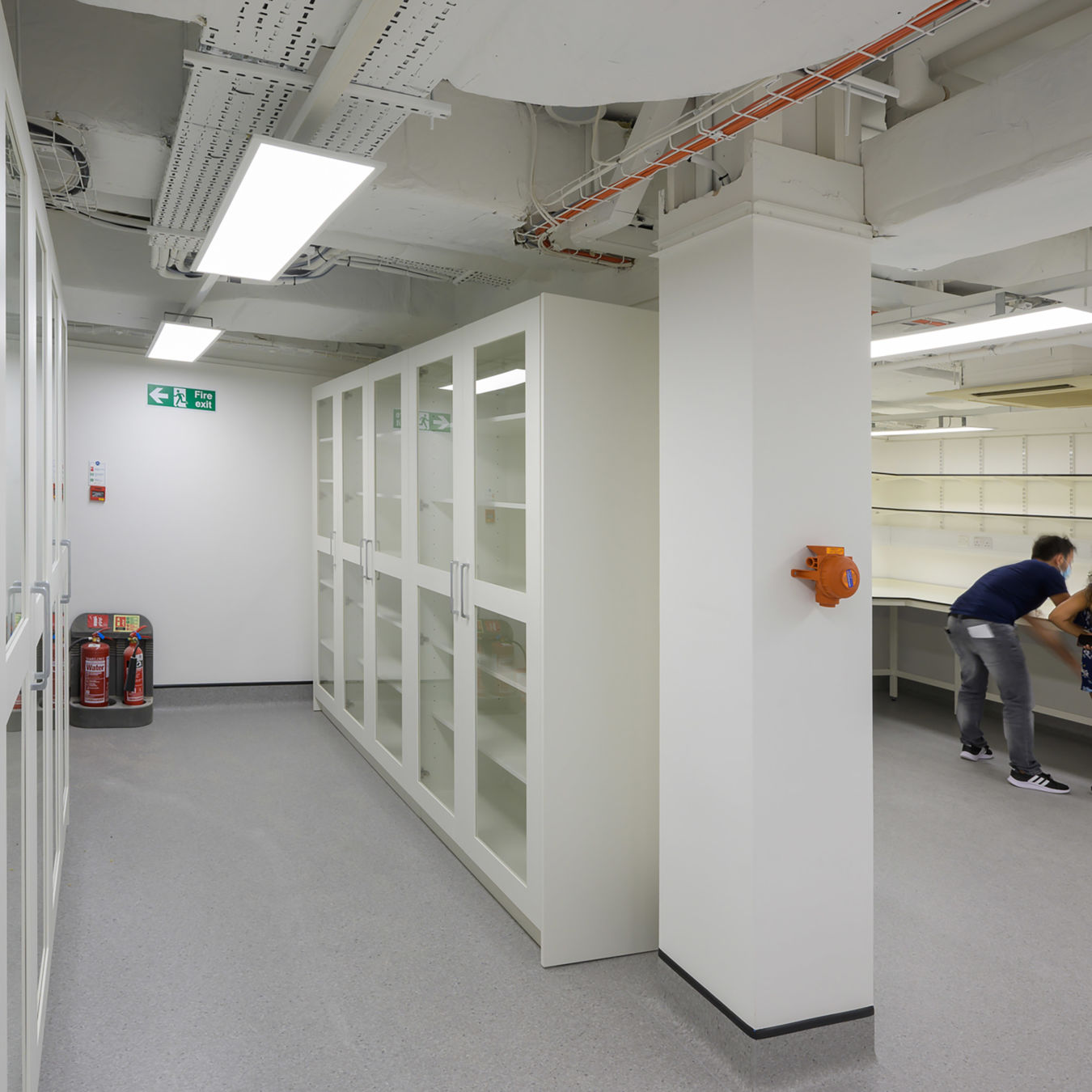 spacious, well-lit laboratory with white cabinets and glass doors. Two people work together at a counter