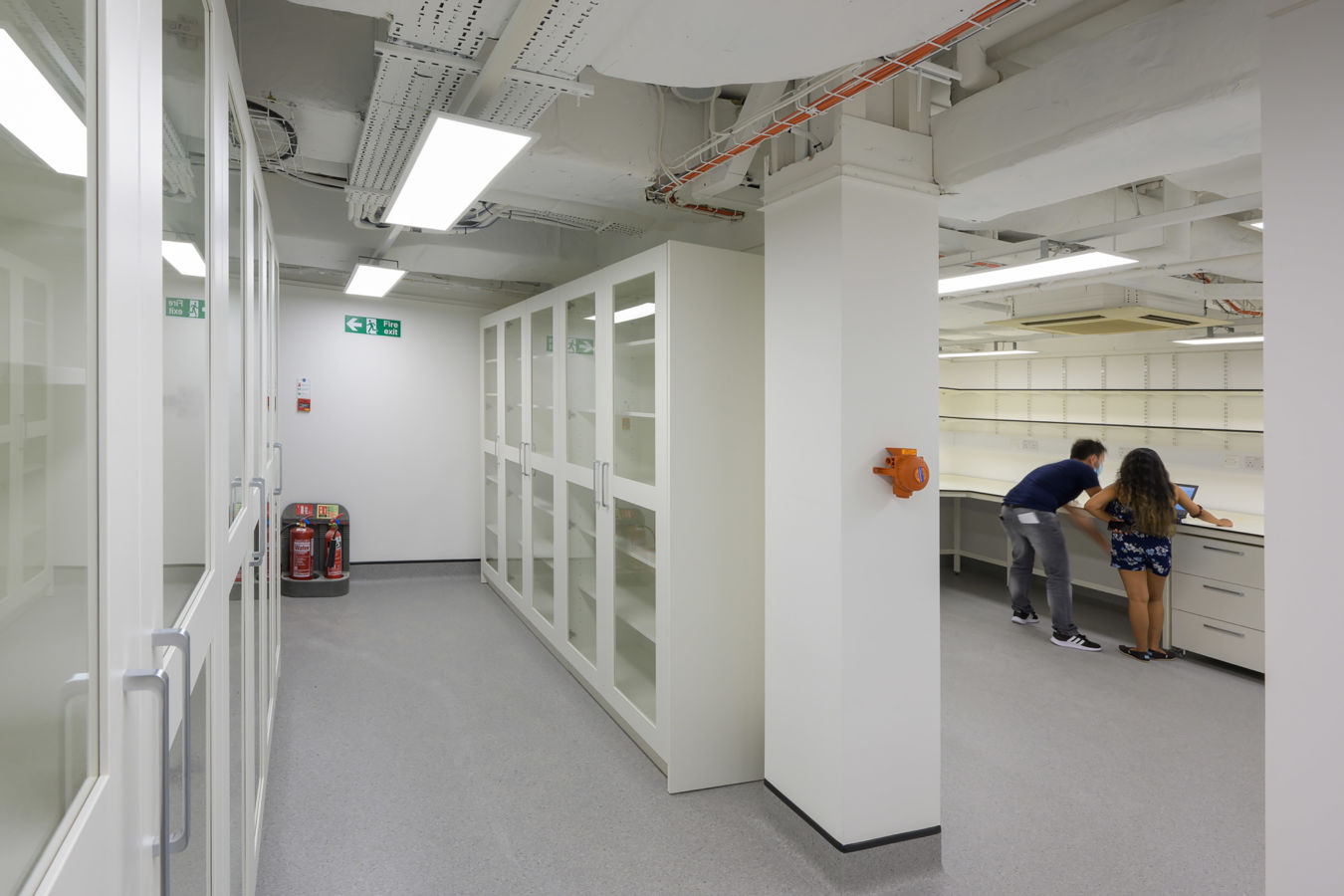spacious, well-lit laboratory with white cabinets and glass doors. Two people work together at a counter