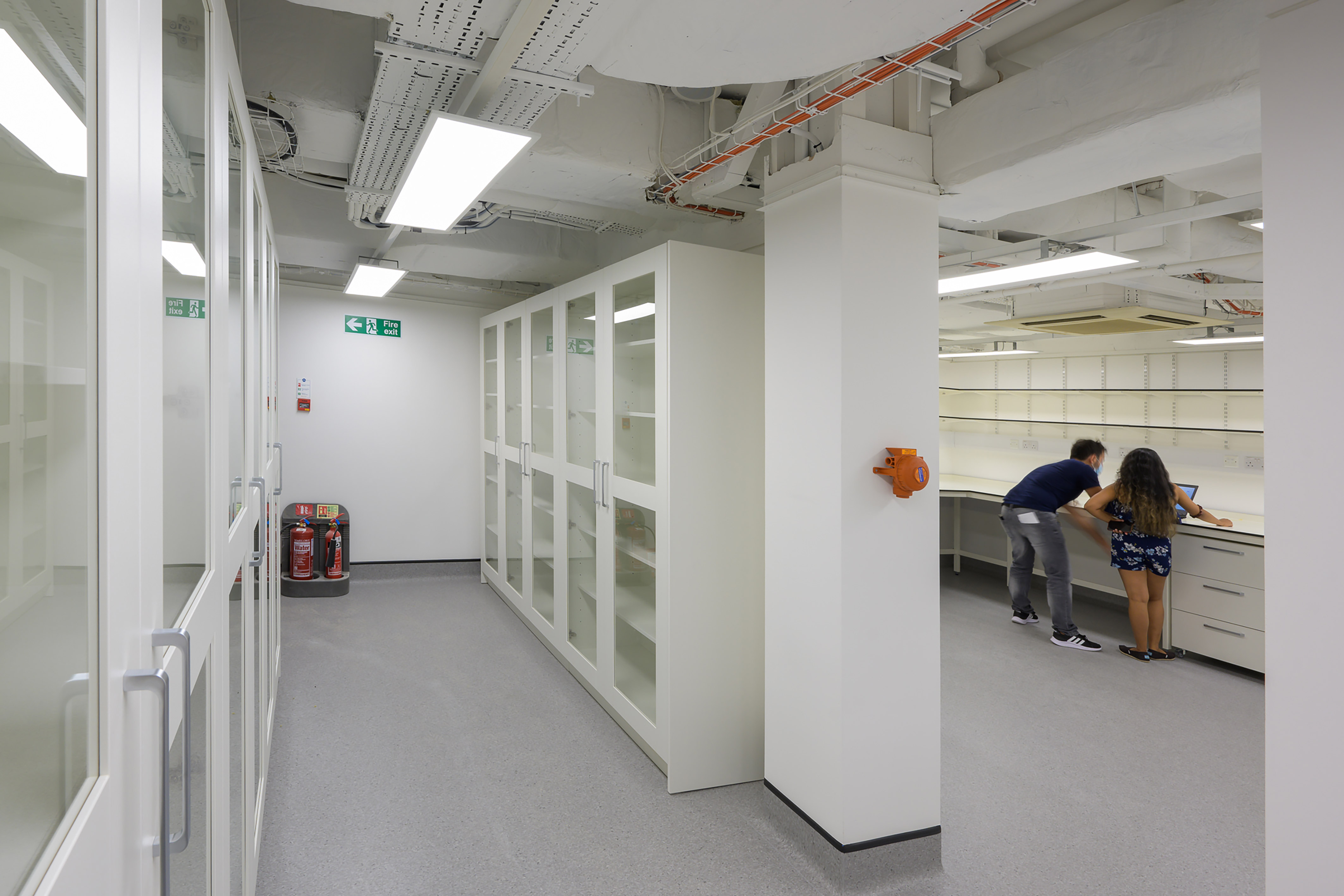 spacious, well-lit laboratory with white cabinets and glass doors. Two people work together at a counter