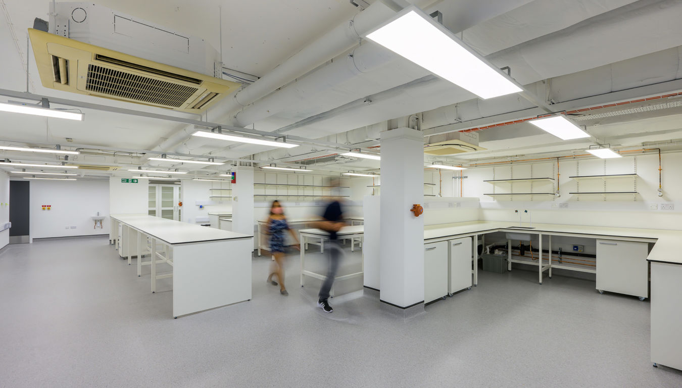 Laboratory with white workbenches and storage shelves, features two blurred figures walking, fluorescent lighting overhead, and a visible air conditioning unit, indicating a spacious, modern scientific environment.